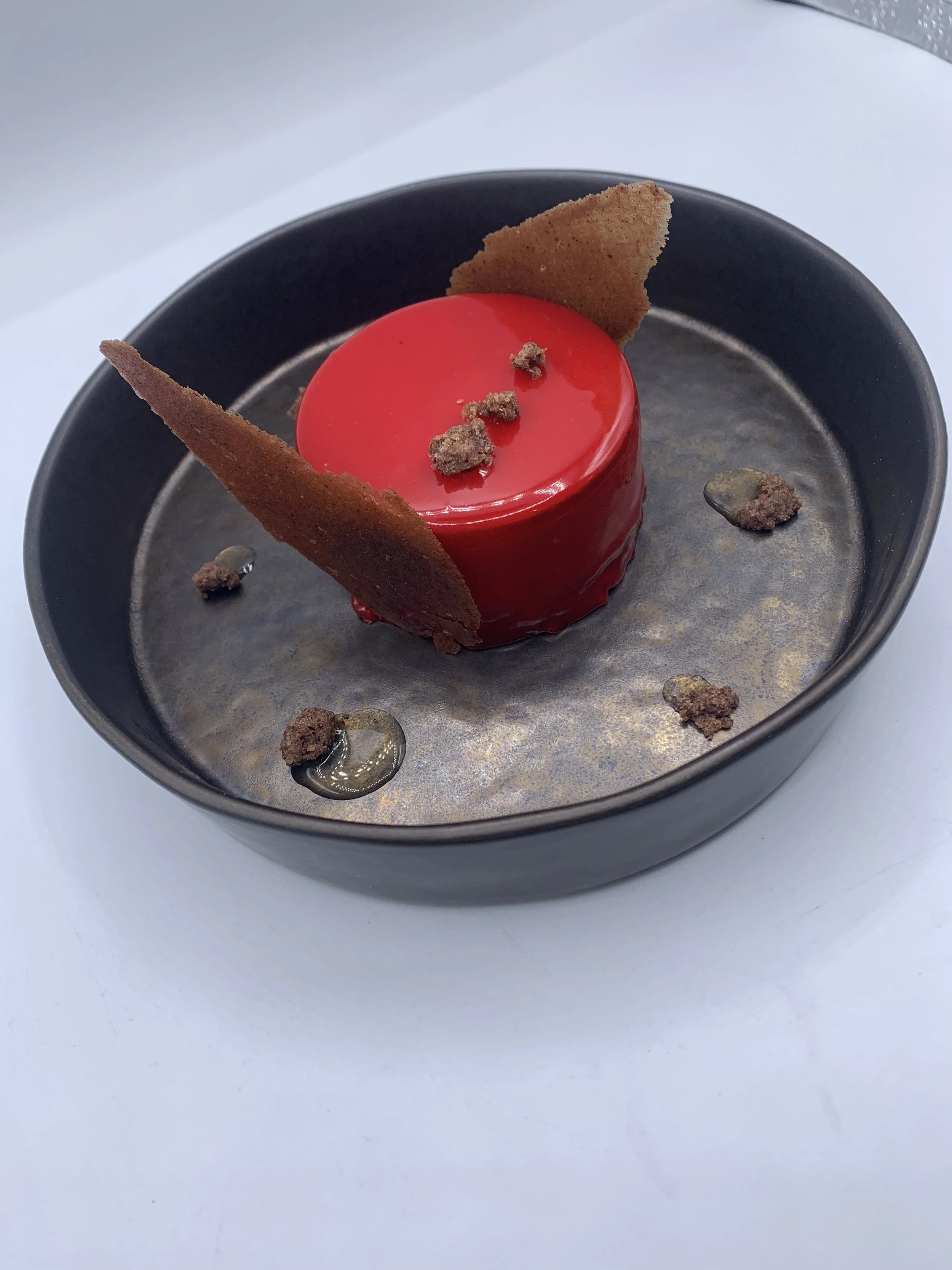 Dessert moderne dans un bol noir, avec un glaçage rouge brillant, décoré de morceaux de biscuit ou de croustillants, et deux fines plaques de biscuit ou de caramel.