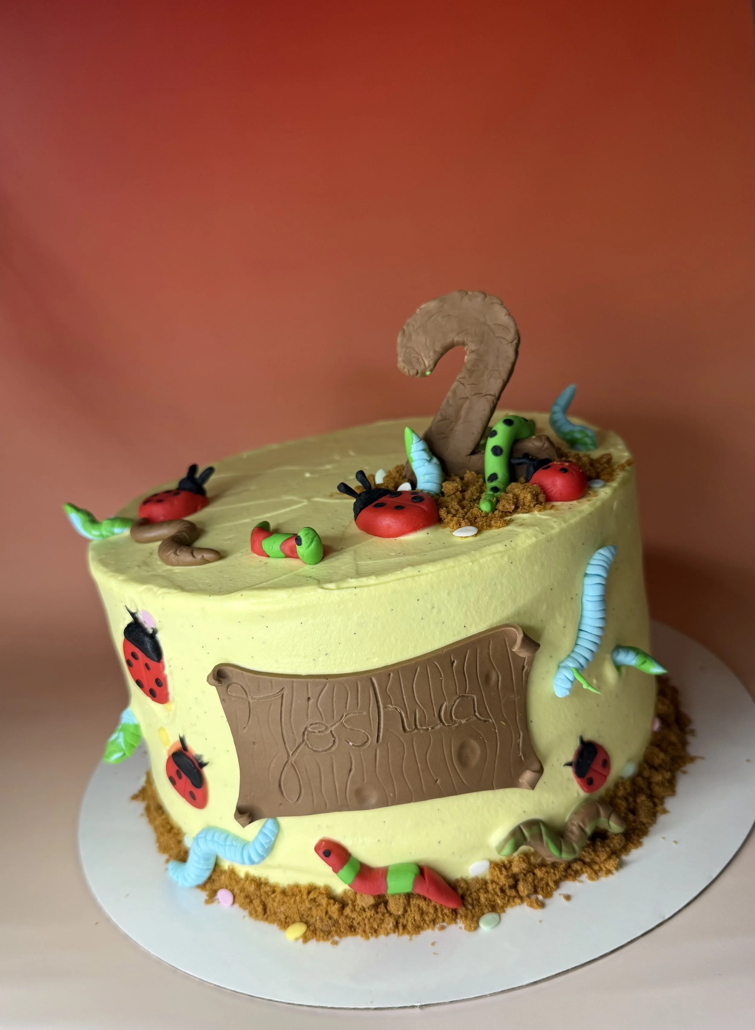 Gâteau d'anniversaire avec décor de insectes en bonbons et glaçage jaune, représentant un environnement de forêt ou de sol, avec un grand chiffre 2 en chocolat au sommet.