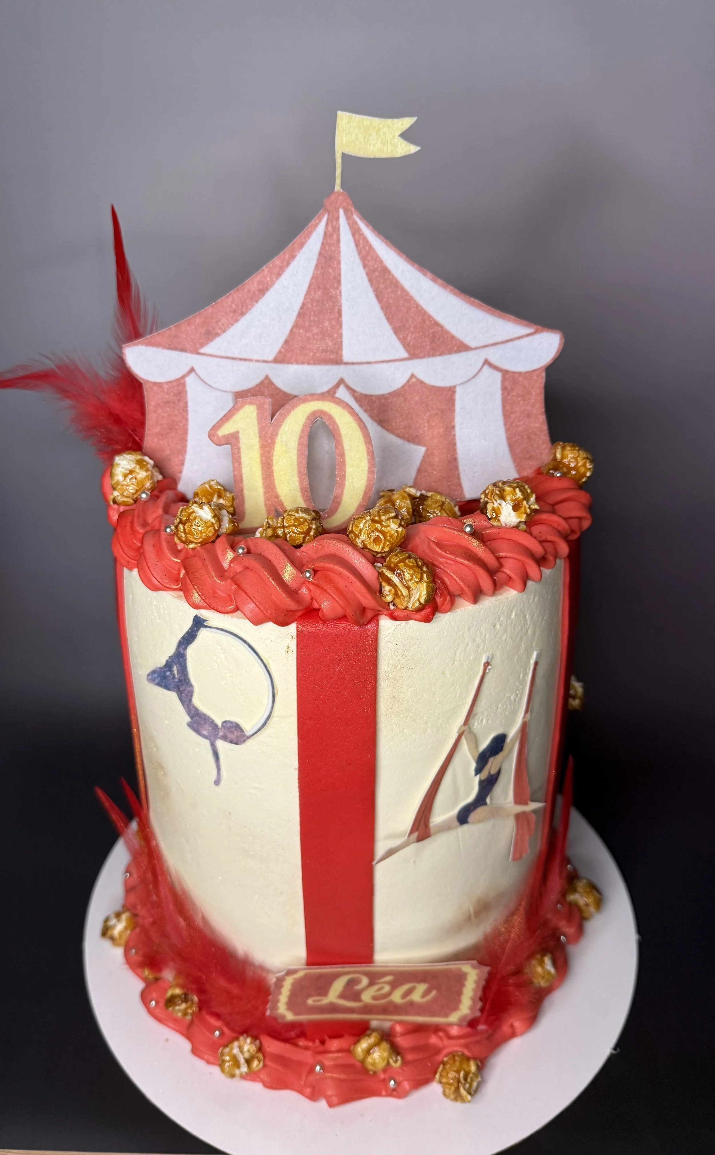 Gâteau d'anniversaire avec un motif de chapiteau de cirque, décoré de caramel au beurre, d'une plume rouge, d'une tête de clown, avec le nom 'Léa' et le chiffre 10 indiquant un anniversaire.