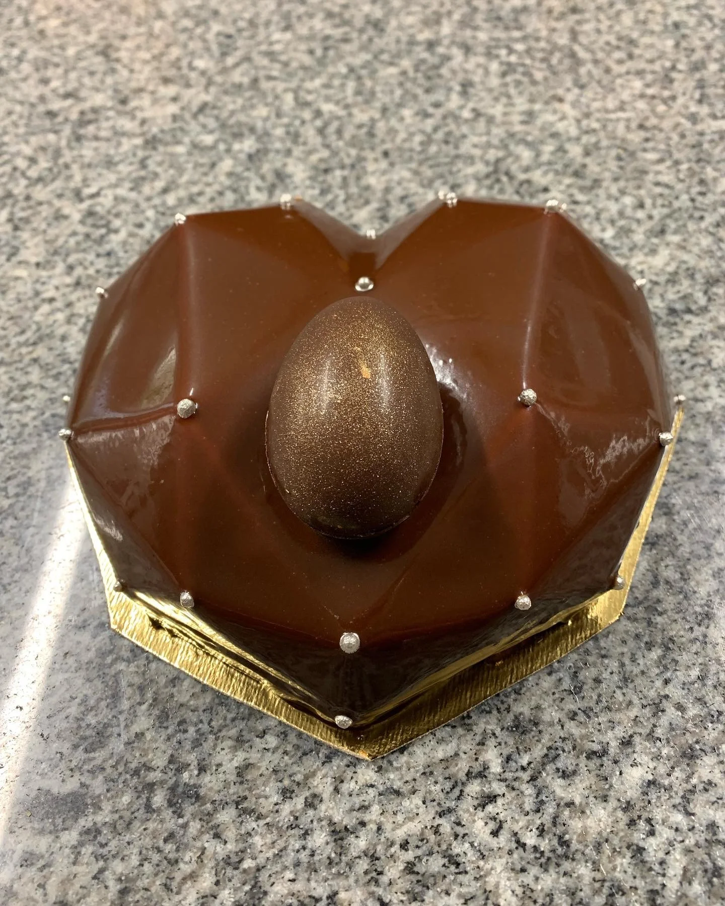 Gâteau en forme de nid d'oiseau avec un œuf en chocolat au centre, décoré de petits perles argentées sur un plateau en or.