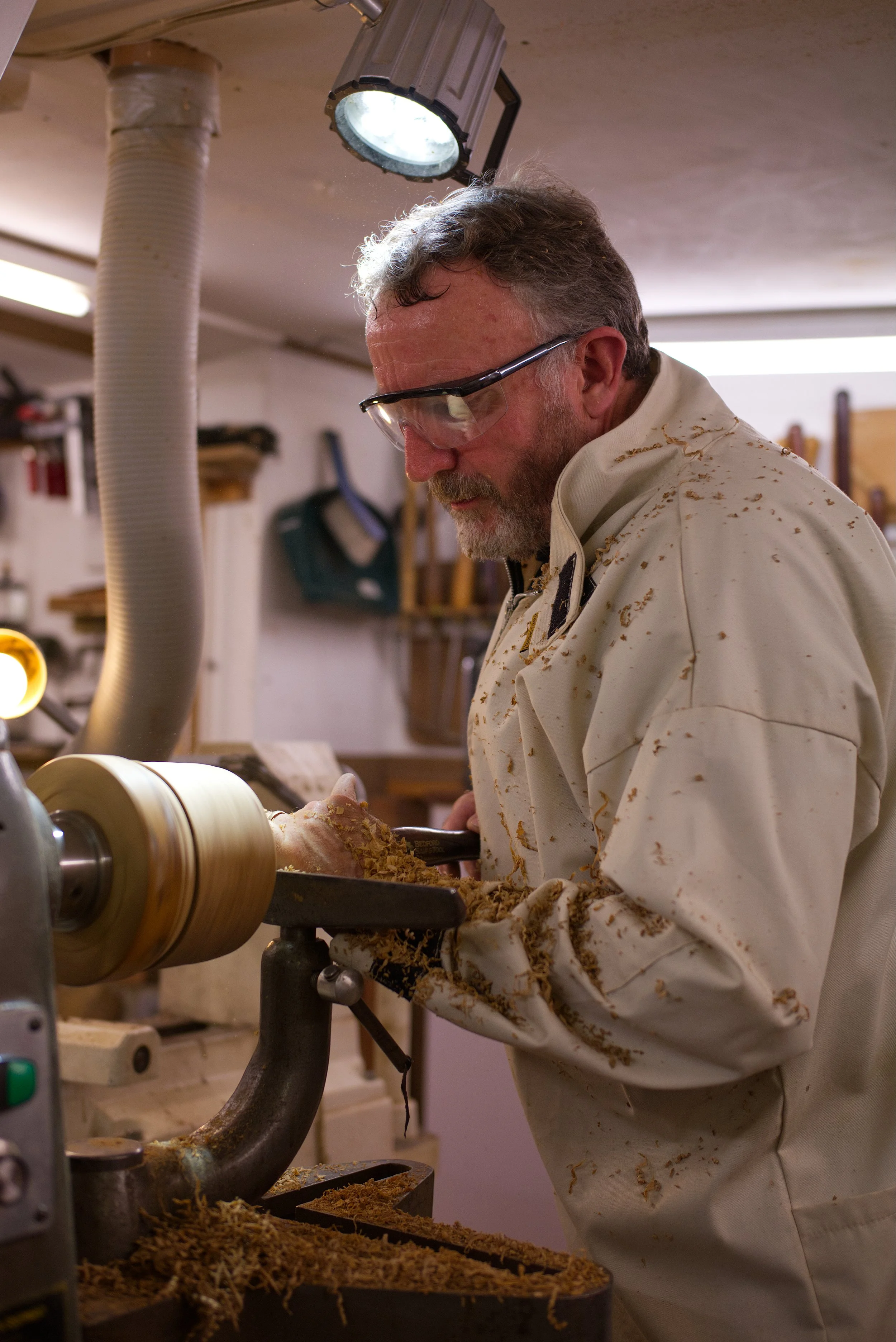 Man with safety goggles and a stained white work coat operating a wood lathe, turning wood shavings and chips in a workshop.