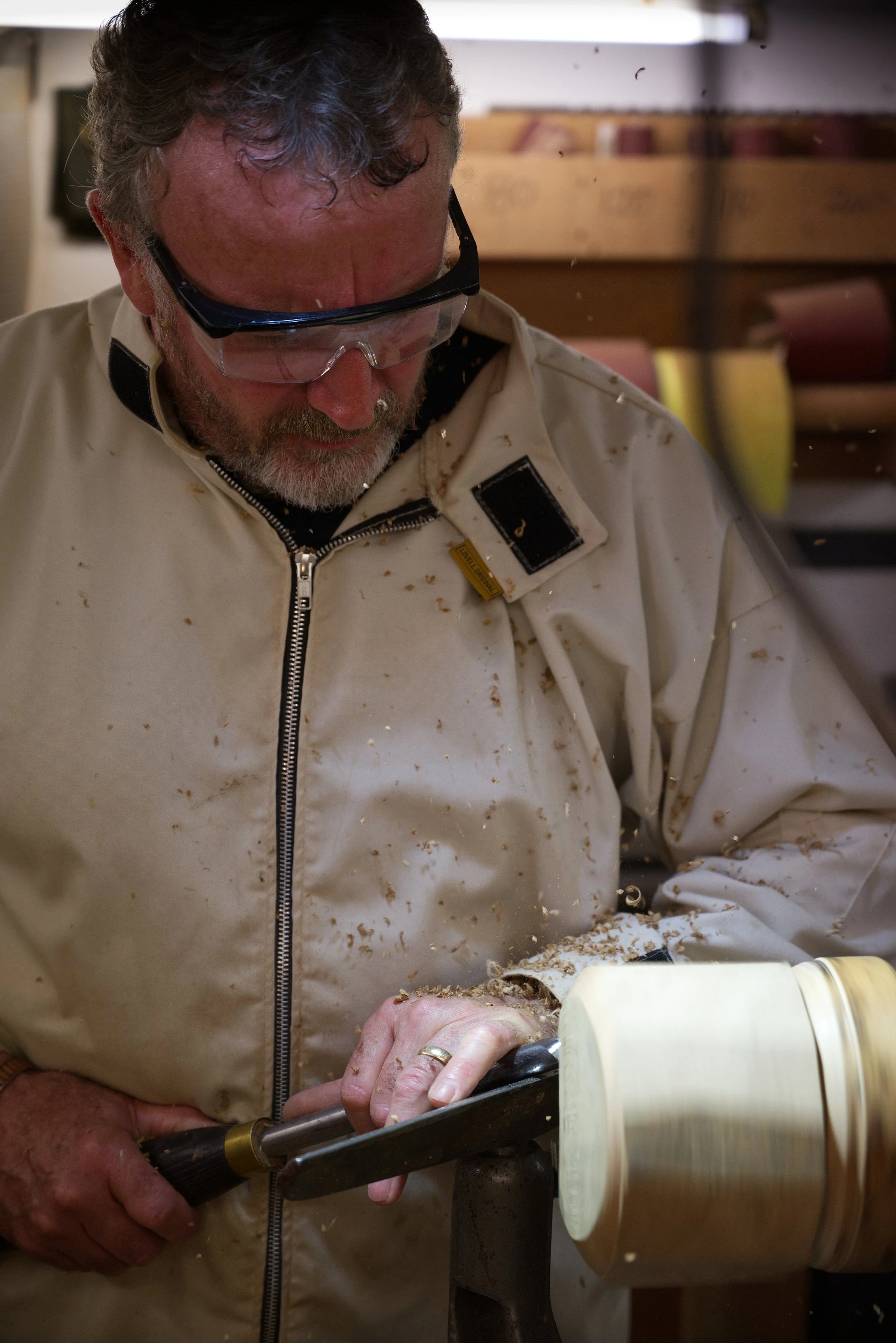 A man wearing safety goggles and a white apron is shaping a piece of wood on a lathe, with wood shavings flying around in a woodworking shop.