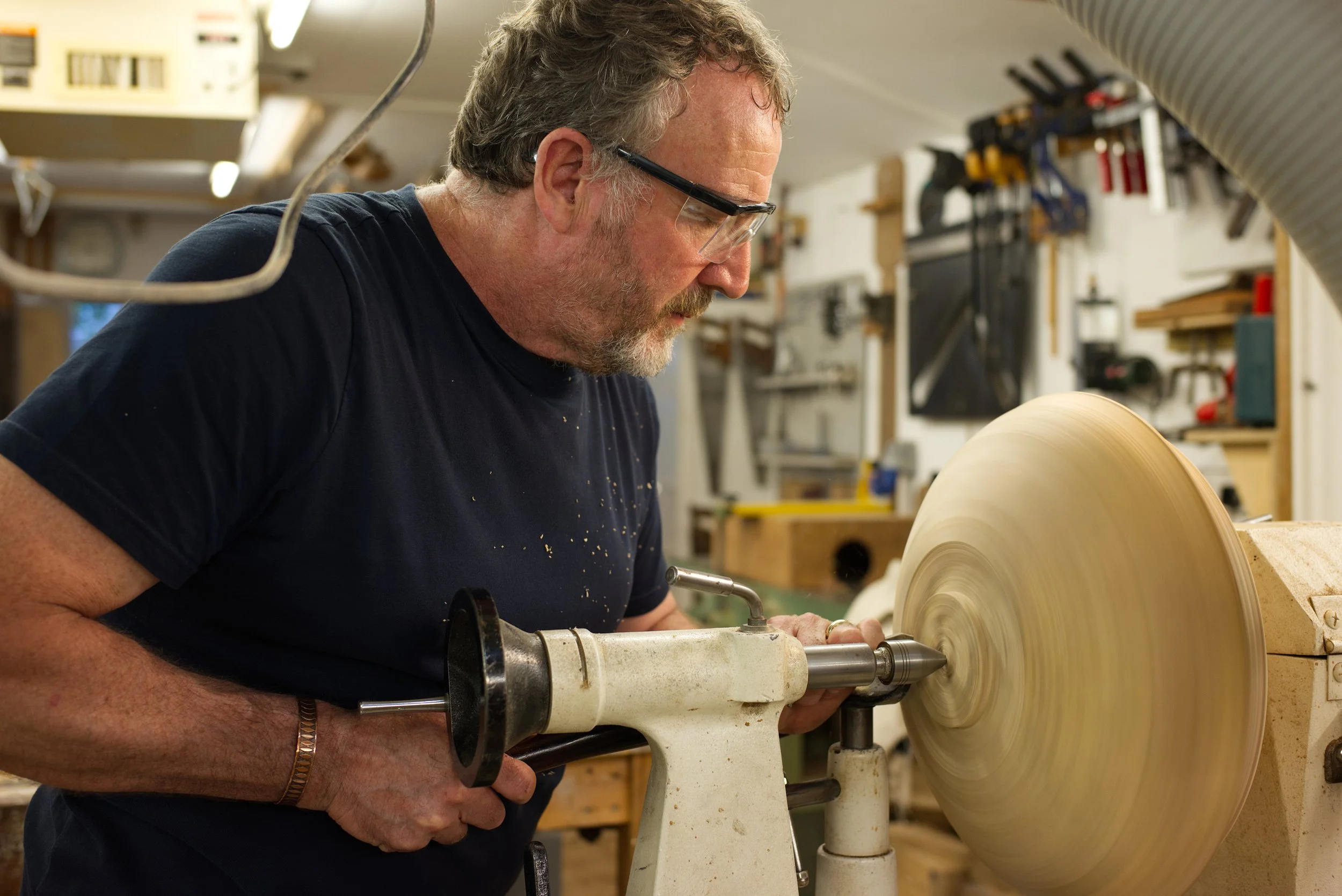 A man wearing safety glasses operates a wood lathe in a woodworking shop, shaping a large piece of wood into a rounded object.