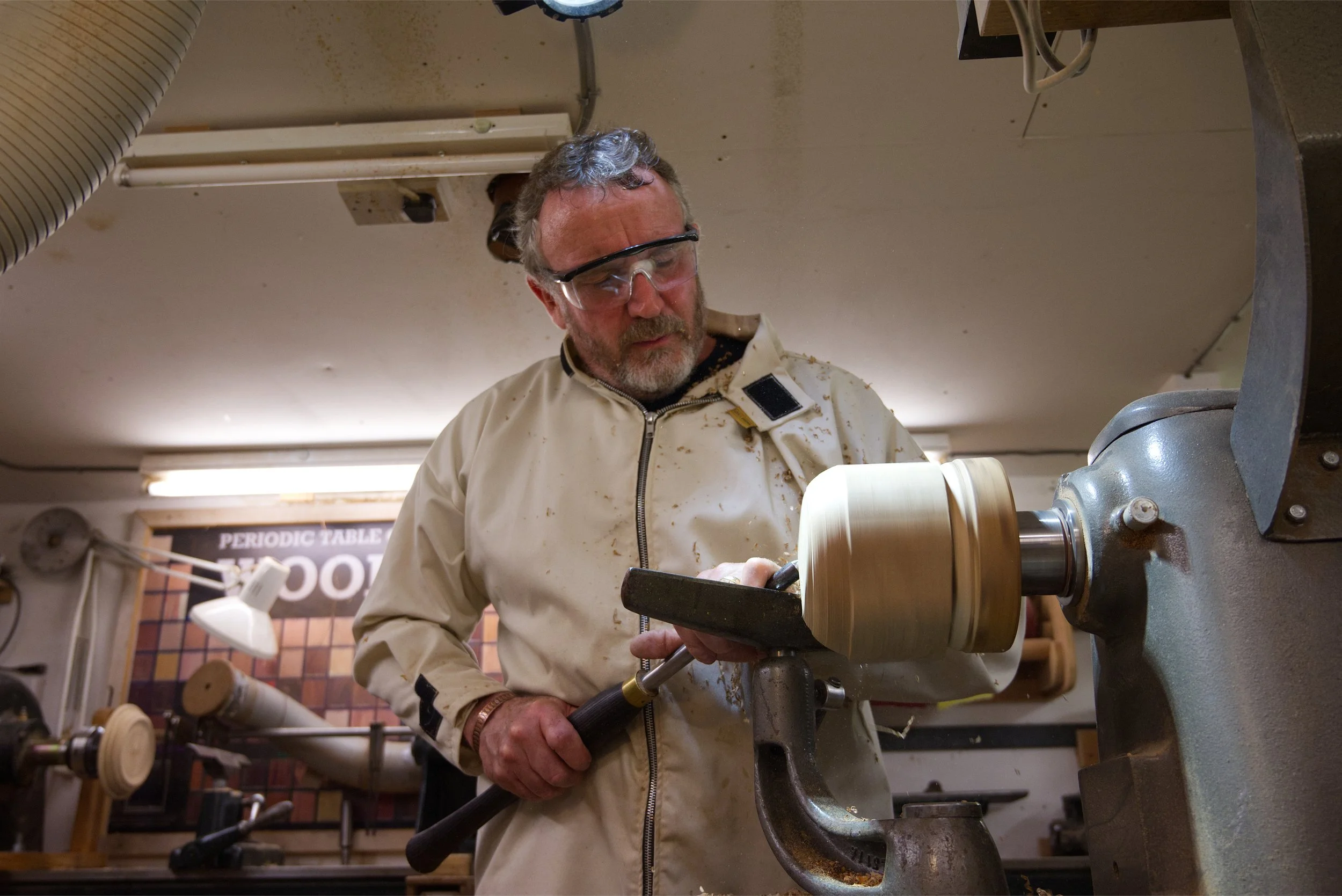 A man with gray hair and a beard wearing safety goggles and a beige jacket working on a woodworking lathe in a workshop.