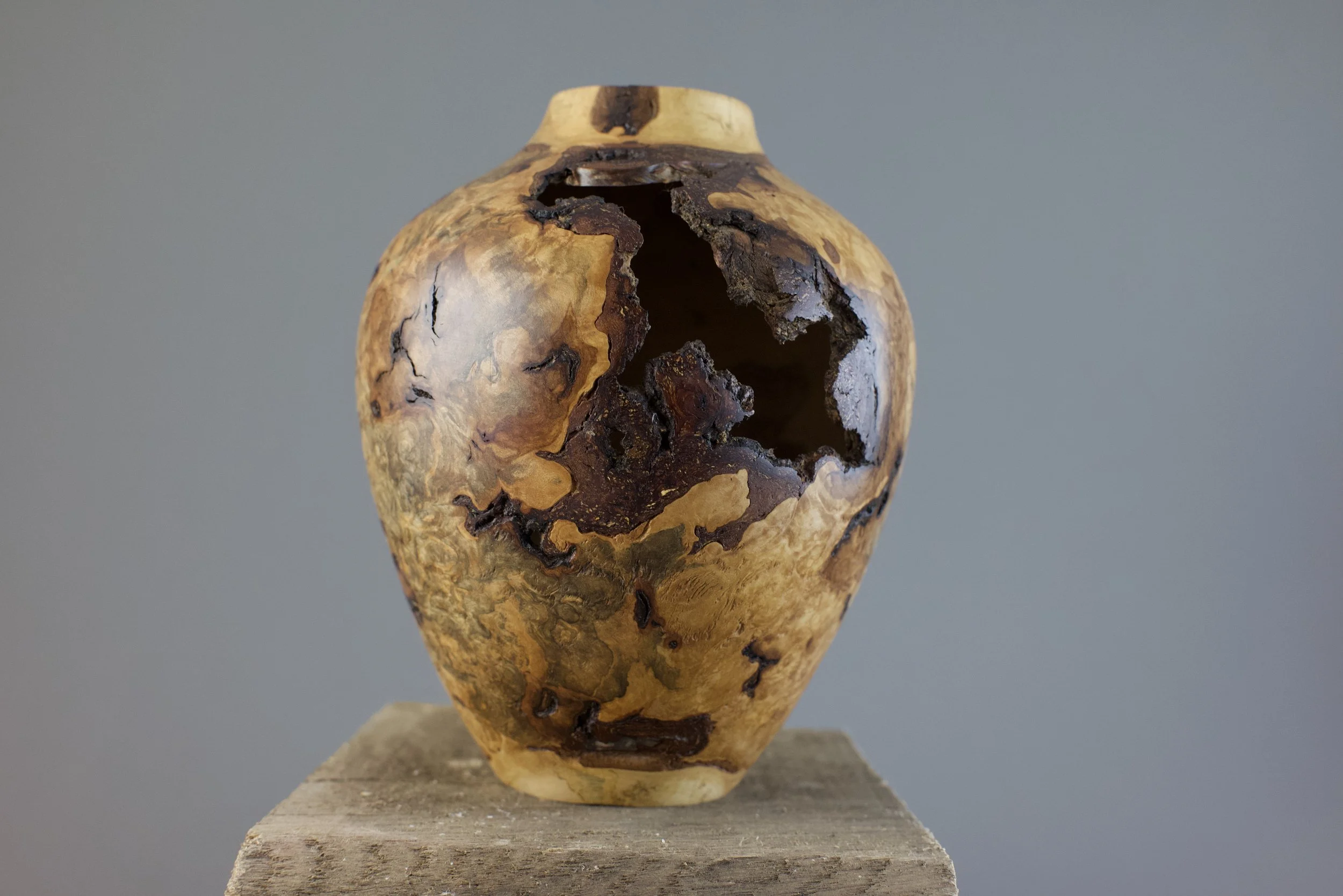 A small, gourd-shaped sculpture with a hollow center and irregular, natural patterns, placed on a wooden block against a plain gray background.