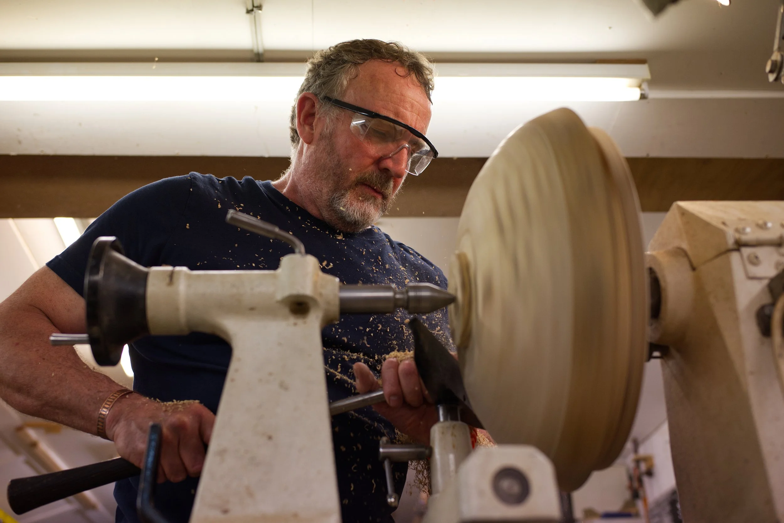 A man wearing safety glasses and a navy blue shirt is working with a lathe in a woodworking workshop. He is focused on shaping a piece of wood, with sawdust visible around him.