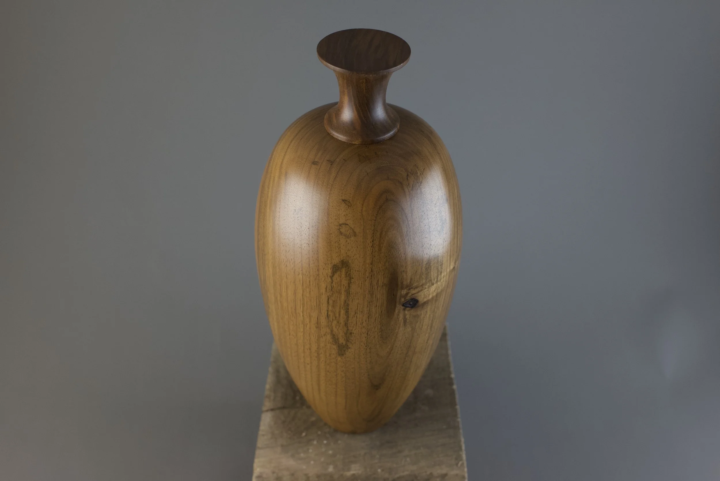 A wooden sculpture resembling an abstract human figure with a rounded top, set against a plain gray background.