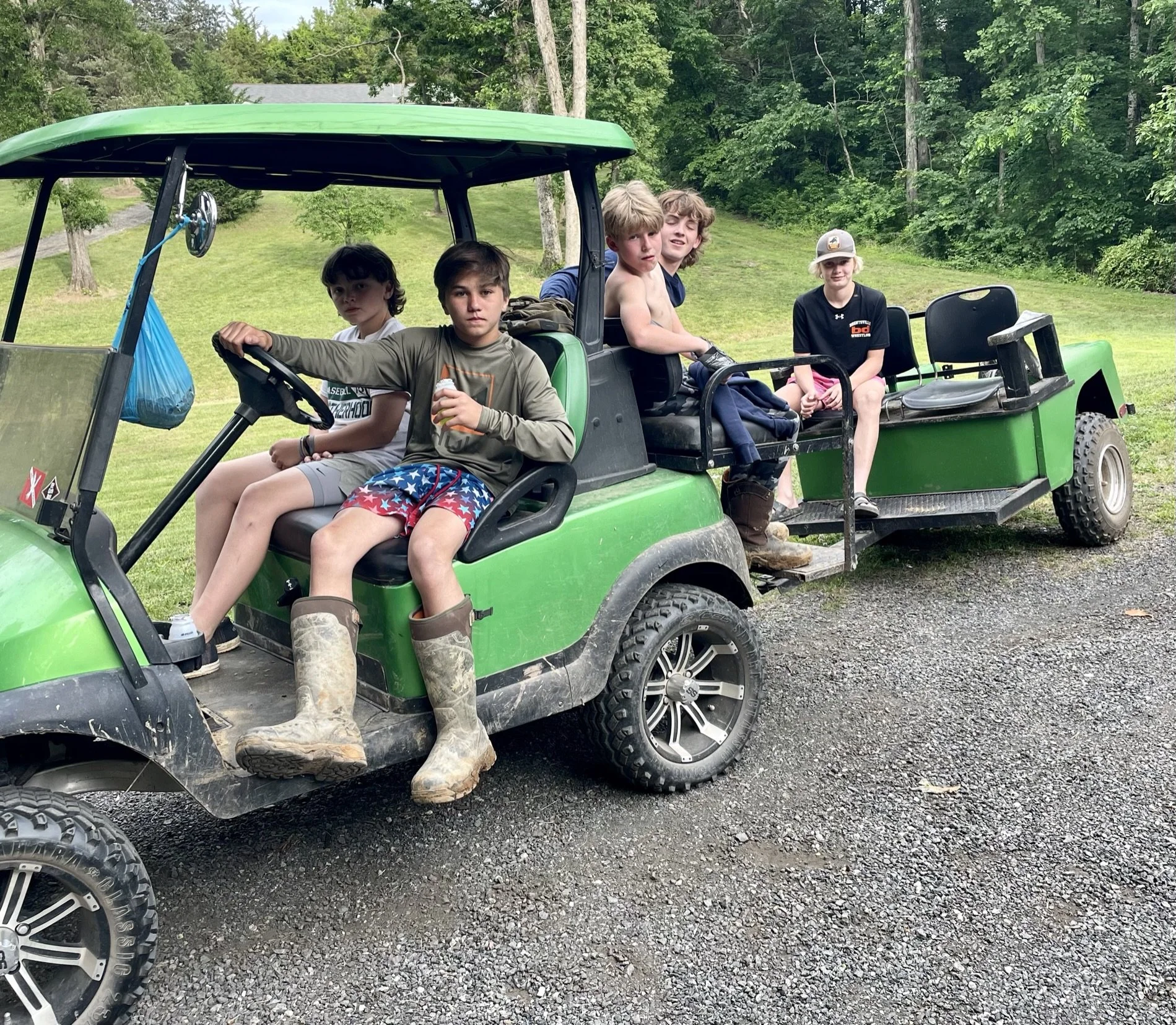 A group of five children sitting in a green golf cart outdoors on a grassy and gravel area, with trees and greenery in the background.