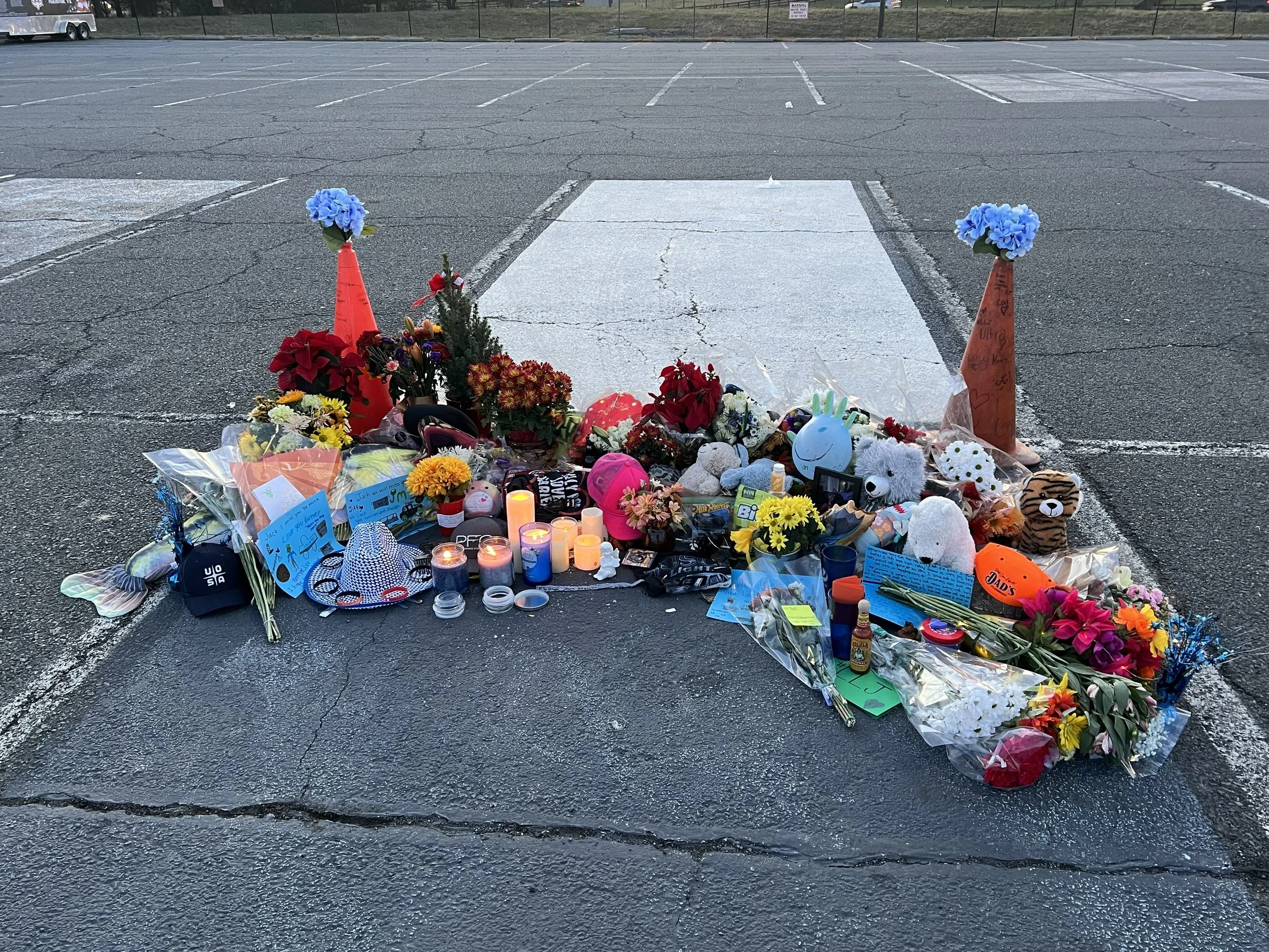 A memorial with flowers, candles, stuffed animals, handwritten notes, and candles placed on the ground in a parking lot.