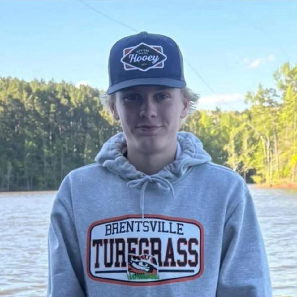 A young person with blonde hair wearing a gray hoodie and a dark baseball cap that says "Hockey" is standing outside near a body of water with trees in the background.