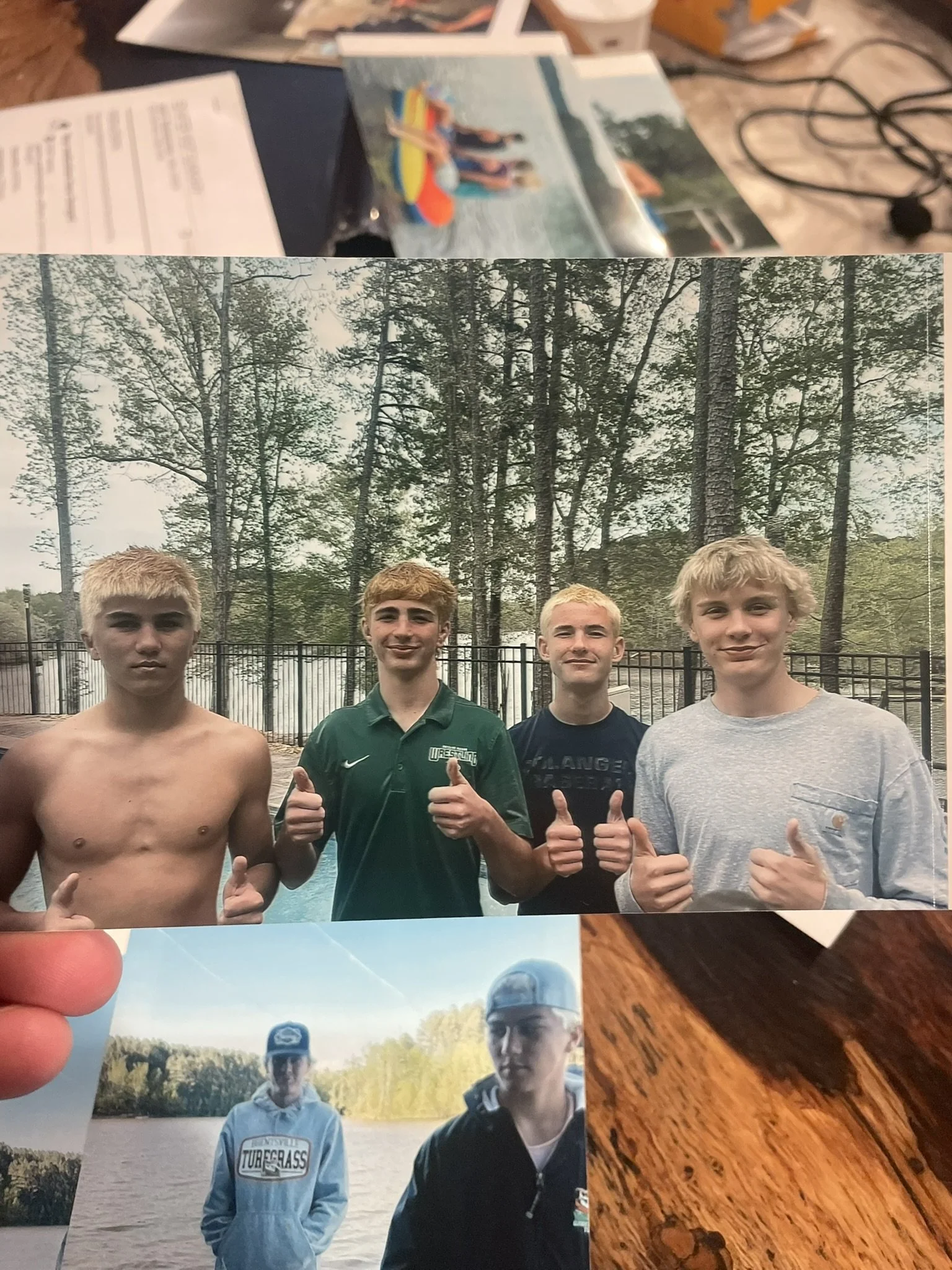 Four young men standing outdoors by a lake with trees in the background, smiling and giving thumbs-up. One is shirtless, others are in hoodies and t-shirts.