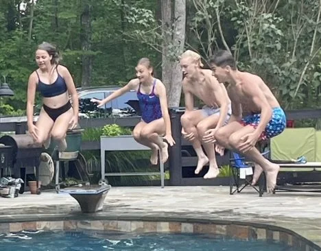 Four young people, two women and two men, are jumping into a swimming pool from a deck surrounded by trees.