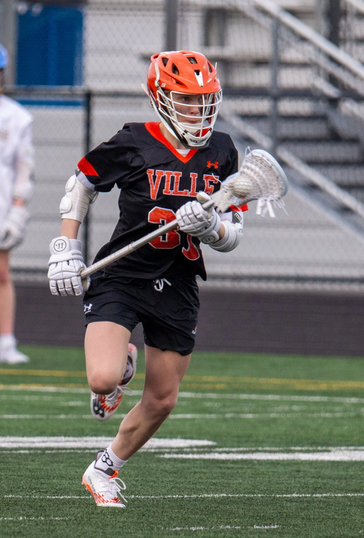 A female lacrosse player wearing a black jersey with 'VILL' in orange letters, orange helmet, white gloves, holding a lacrosse stick, running on a green field with white yard lines.