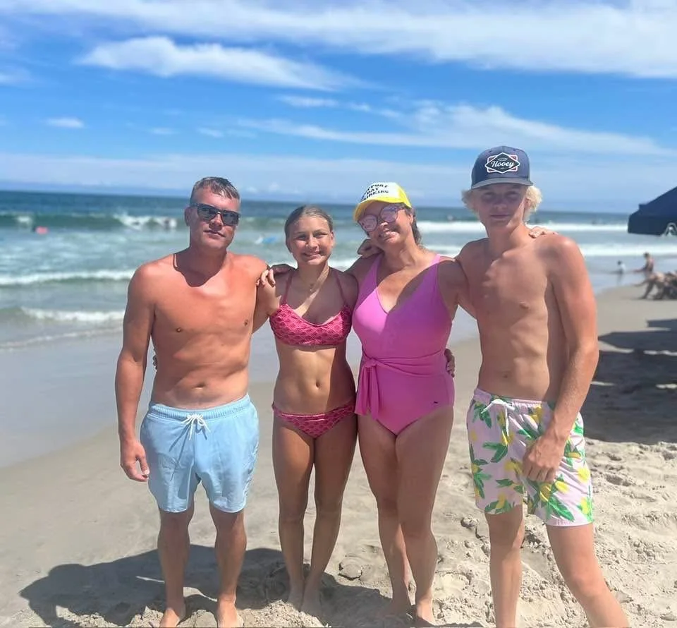 Four people standing on a sandy beach next to the ocean, posing for a photo. They are dressed in swimwear, and there are blue skies with some clouds in the background.