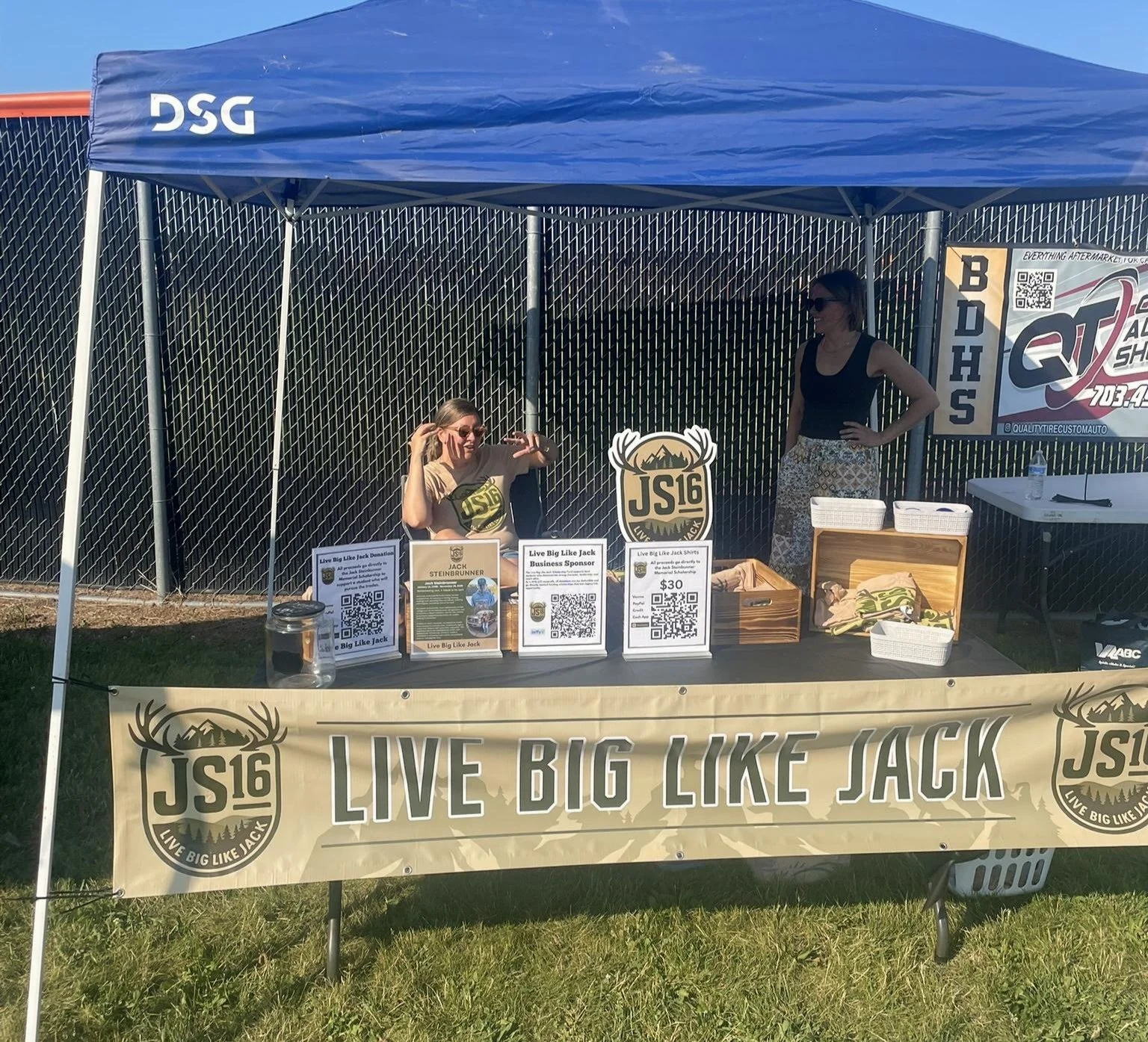 A booth at an outdoor event with a blue canopy tent showing the text "DSG". Two women are behind the booth; one is sitting and making a hand gesture, and the other is standing with her hands on her hips. The booth has signs promoting the 'Live Big Like Jack' event, and various items including T-shirts, baskets, and a water bottle are on the table.