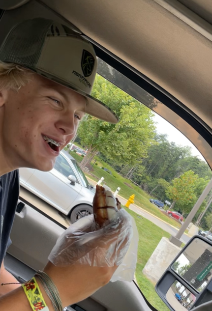 A young man sitting in a vehicle holding a cupcake with a lit candle, smiling and looking at it. Outside the vehicle, there are trees and parked cars.