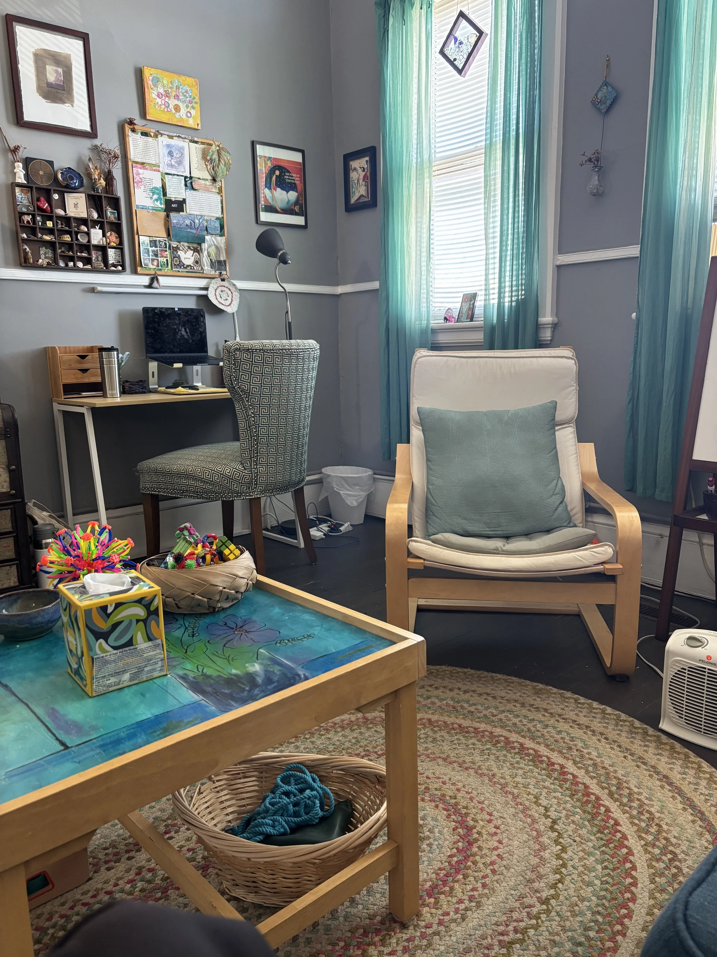 A cozy room with light blue walls, teal curtains, and a window with blinds. The room has a white cushioned chair with a pillow, a small desk with a patterned chair, and wall decorations including framed pictures and a corkboard. There is a colorful t