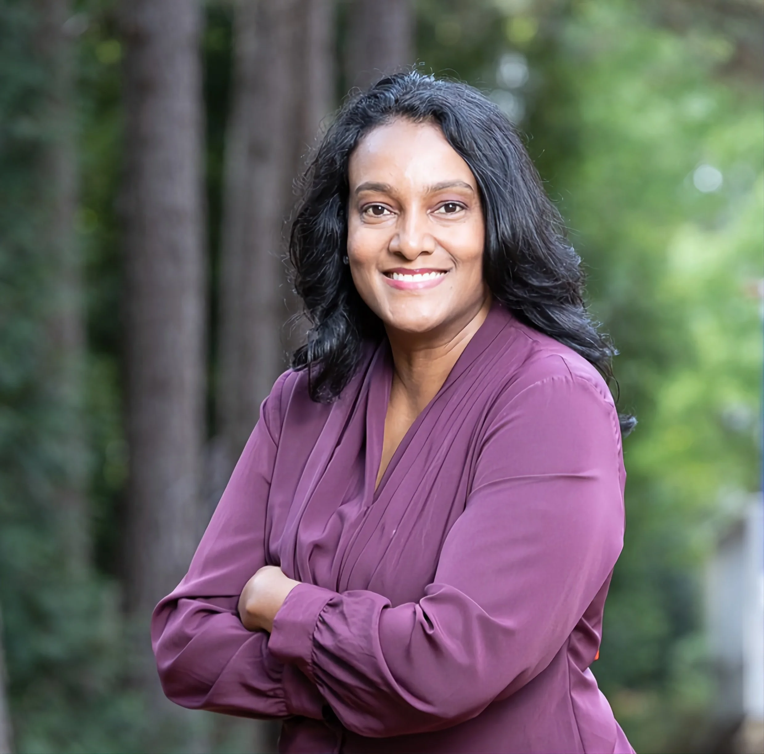 Portrait of a woman with black hair, smiling, wearing a purple blouse, standing outdoors with green trees in the background.
