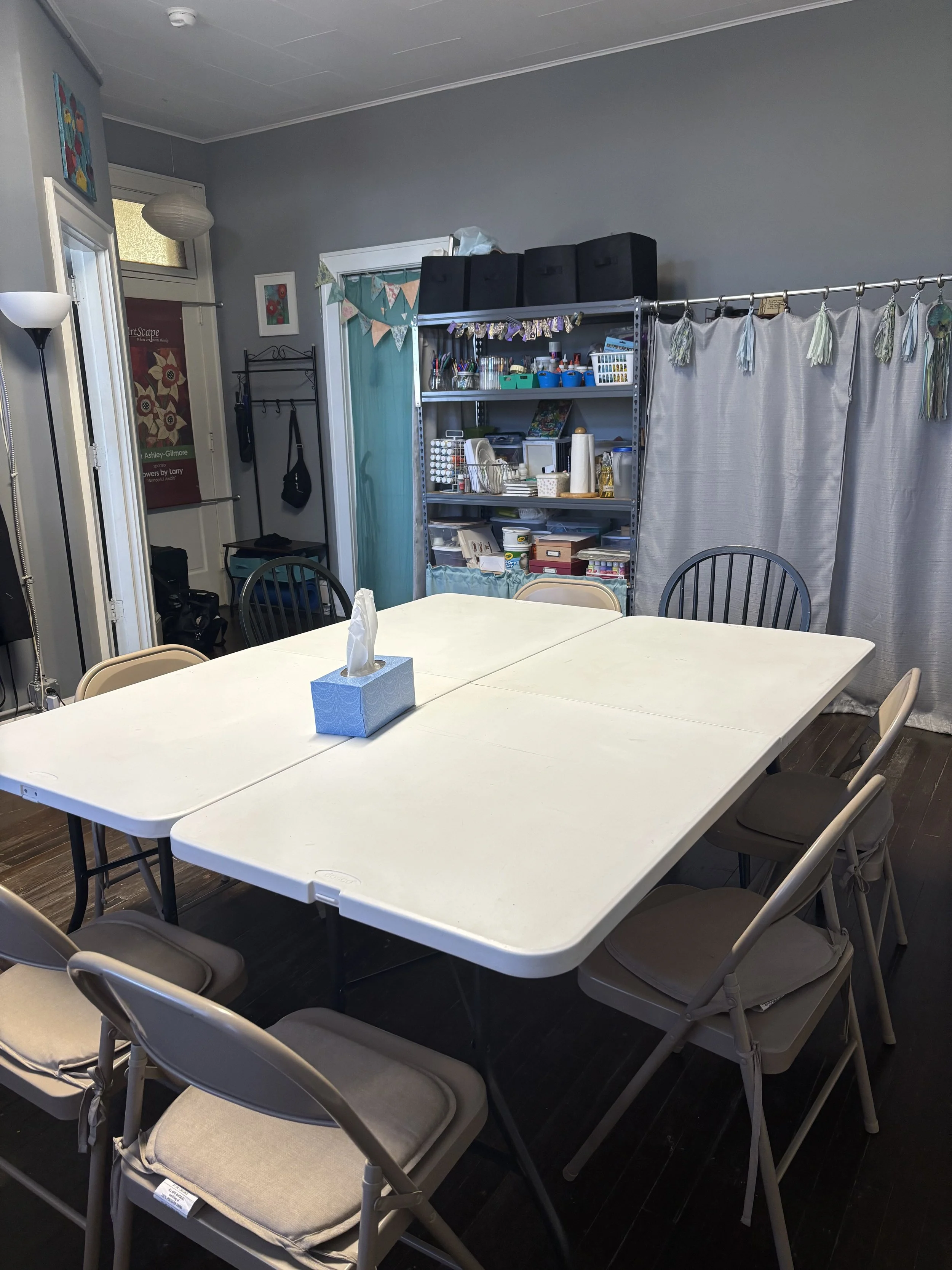 A dining room with a white folding table surrounded by beige and black chairs. On the table, there is a tissue box. In the background, a metal shelf holds various craft supplies, and the room has gray walls with a window, curtains, and decorative ban