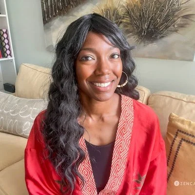 A woman with long, wavy black hair, wearing a red satin robe with gold embroidery on the collar, smiling while sitting on a beige sofa in a living room.