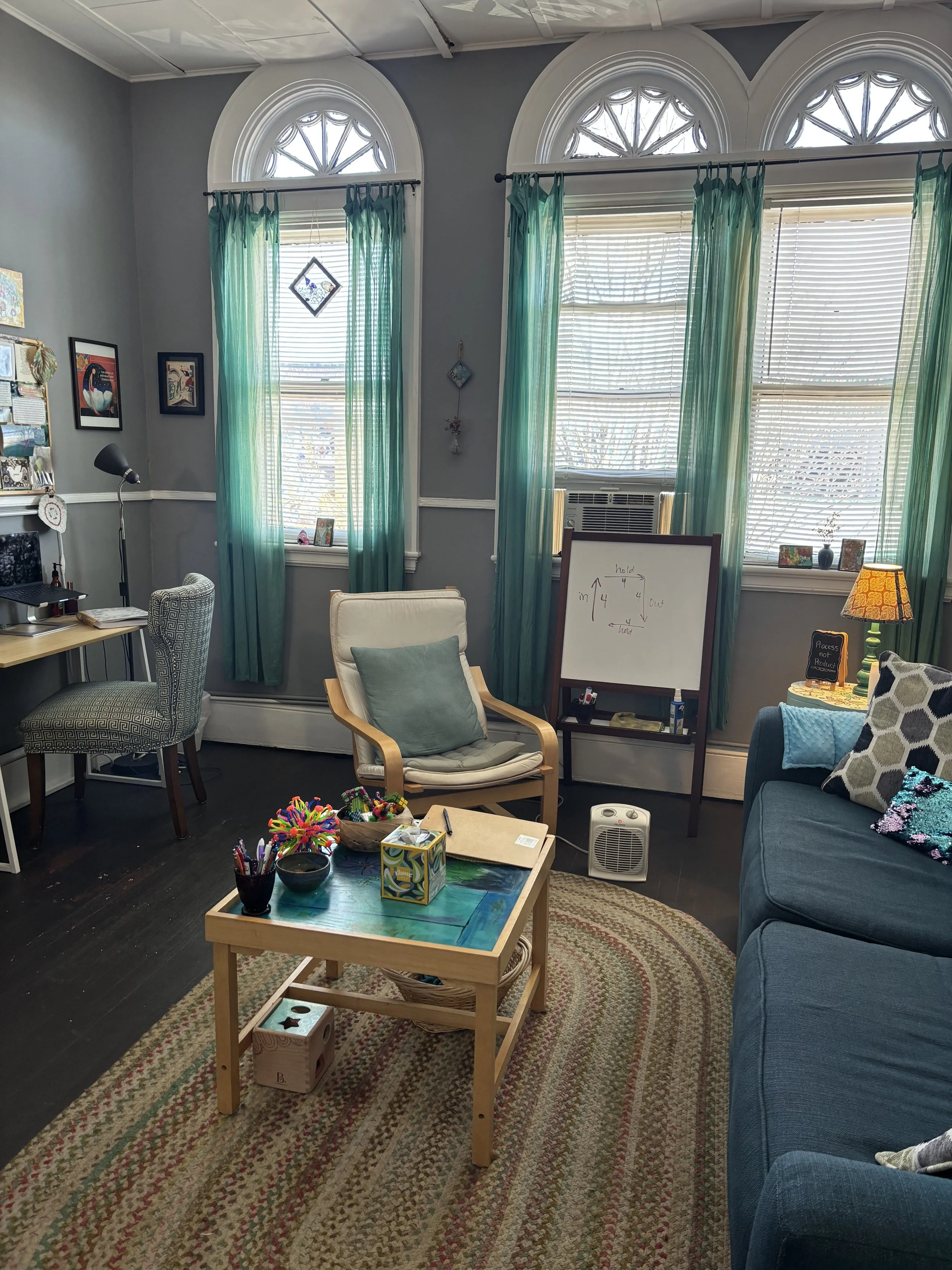 Living room with three tall, arched windows with green curtains, a blue sofa with decorative pillows, a white chair with a pillow, a small table with art supplies, a whiteboard with writing, a wooden side table with a lamp, a desk with a chair, and v