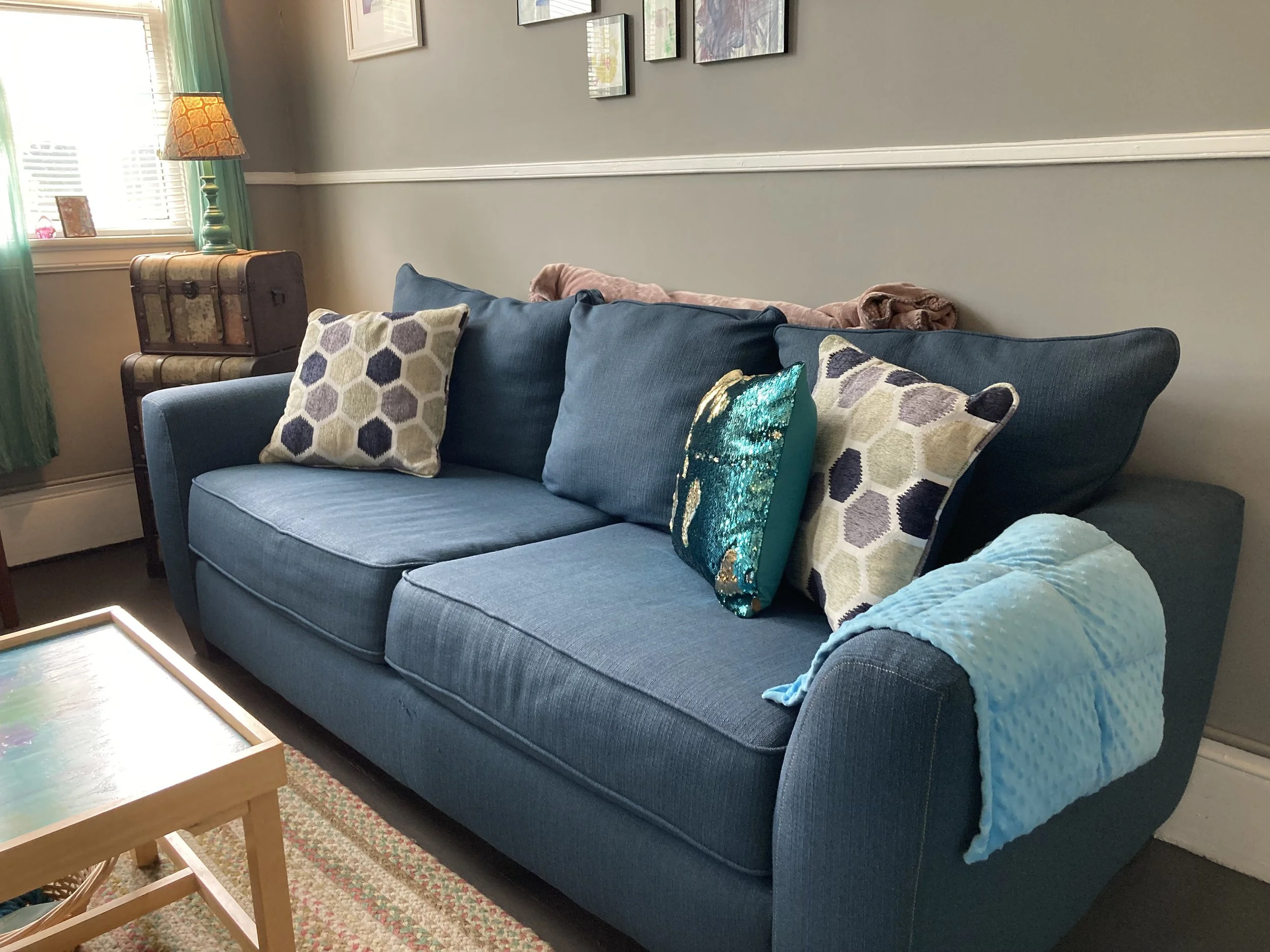 A blue fabric sofa with three cushions, two patterned and one sequined, situated in a brightly lit living room, with a blanket draped over the armrest.