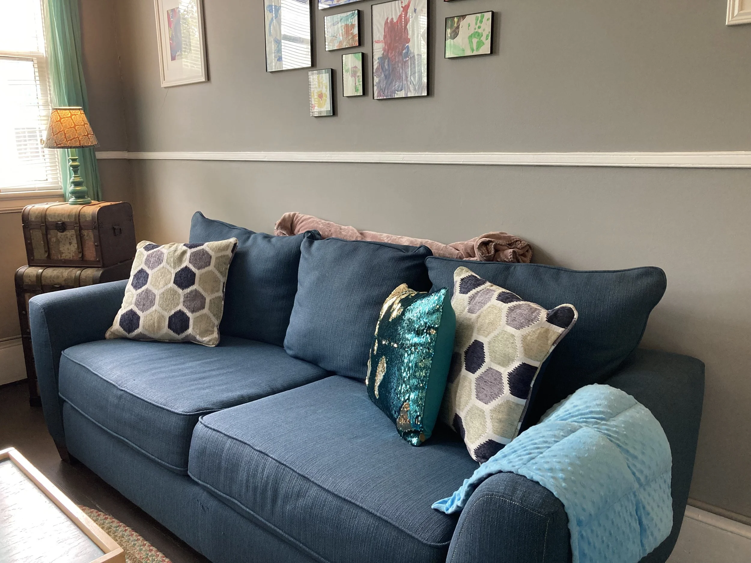 A blue sofa with four pillows, a blanket, and a wooden side table with a vintage trunk underneath a window with green curtains. A lamp is on the side table and a wall with framed, colorful artwork.