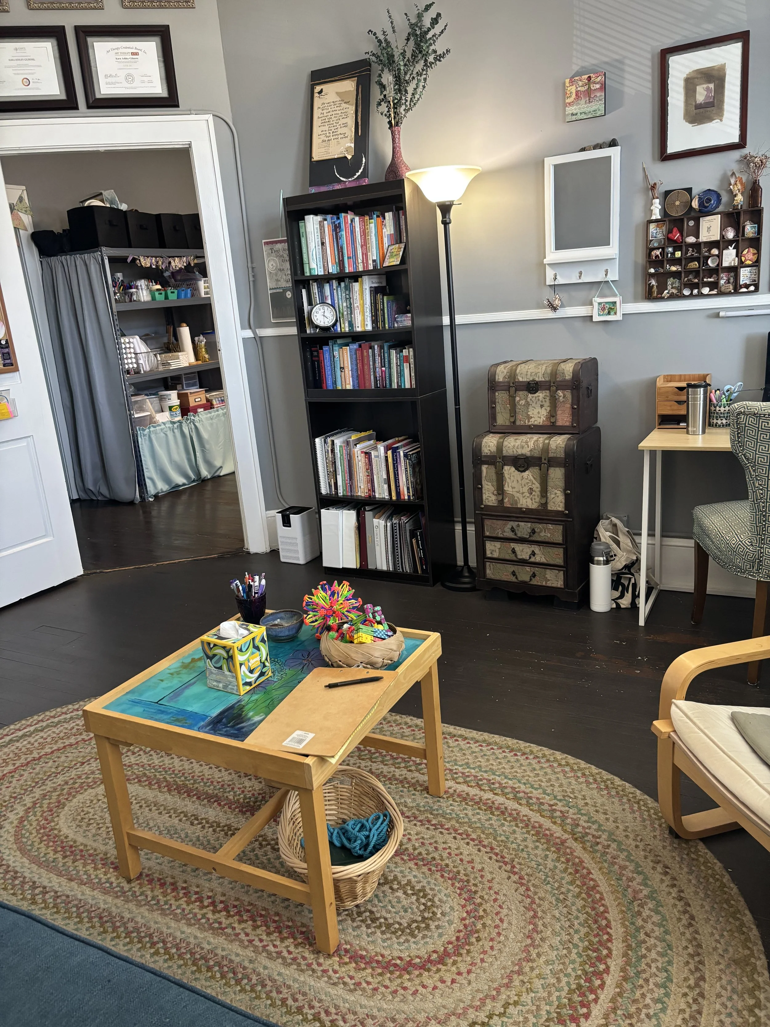 A cozy living room with a wooden coffee table on a multicolored braided rug, a black bookshelf filled with books, and various decorations on the wall. A tall lamp and a small chest of drawers are also visible.