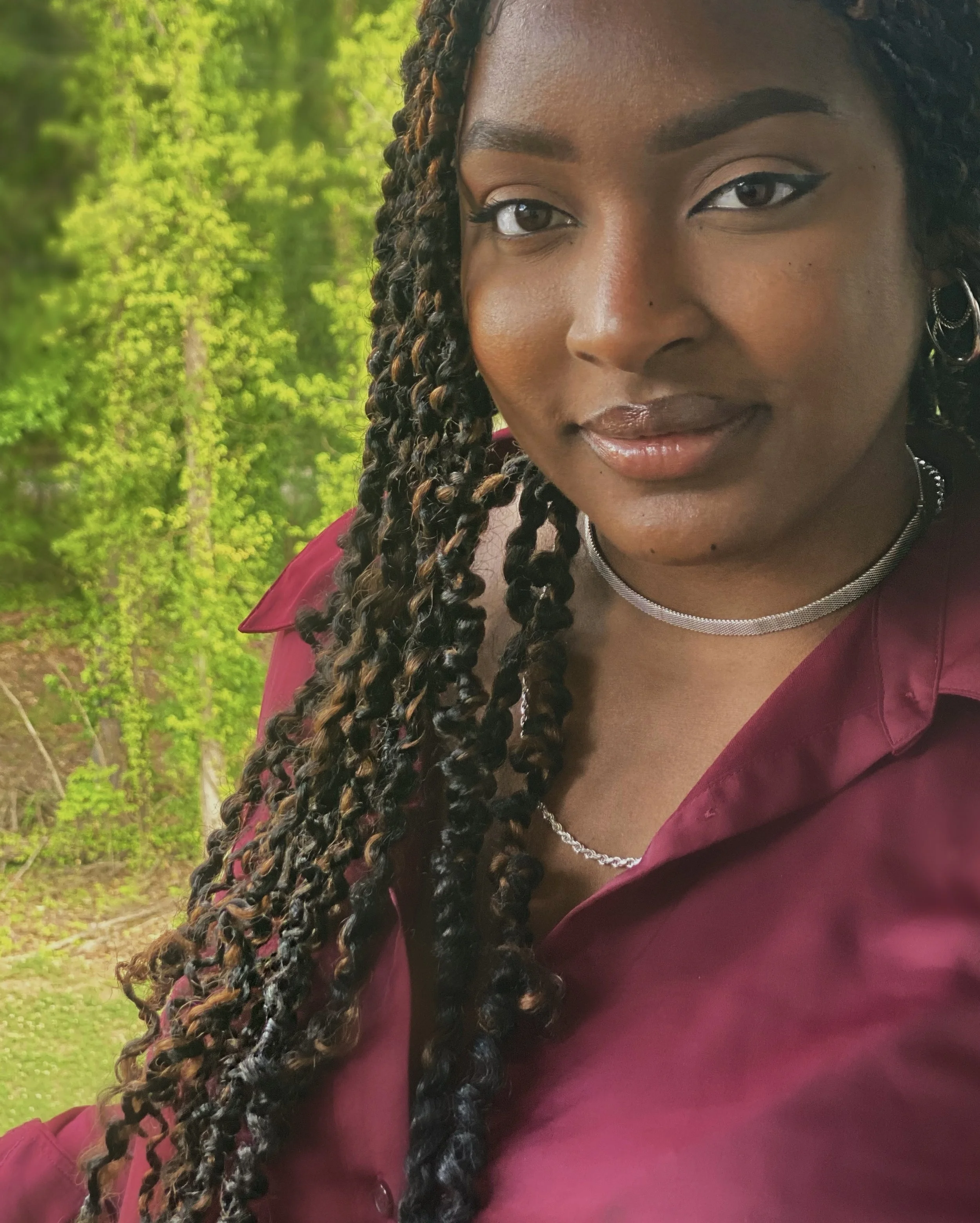 A woman with dark curly hair styled in twists, wearing a maroon shirt, silver choker, and earrings, outdoors with green foliage in the background.