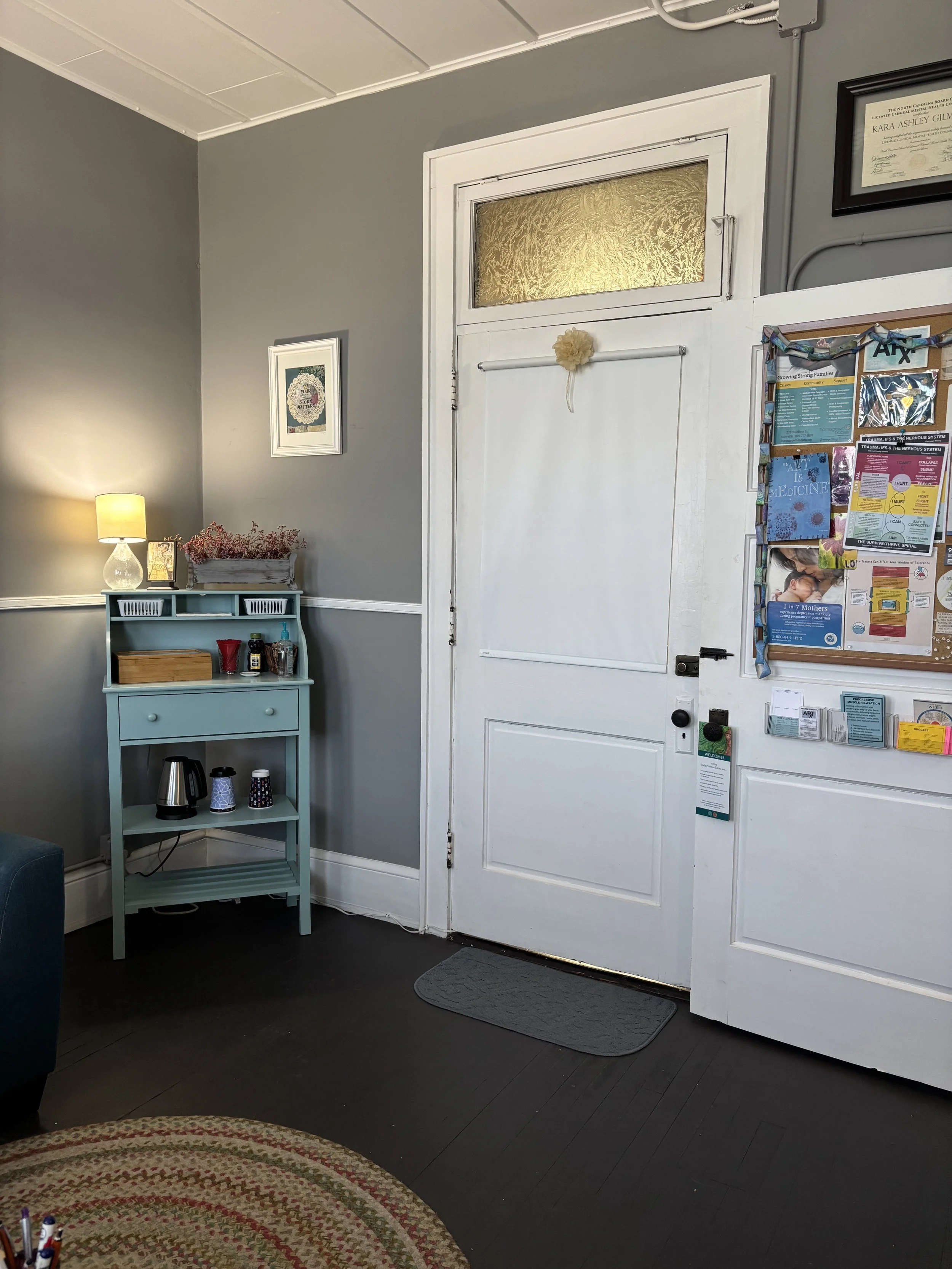 Interior of a waiting room or office space with a white door, gray walls, a small blue shelving unit with cups and a kettle, a wall-mounted bulletin board with papers, a framed certificate, a small lamp, and a rug on dark hardwood floors.