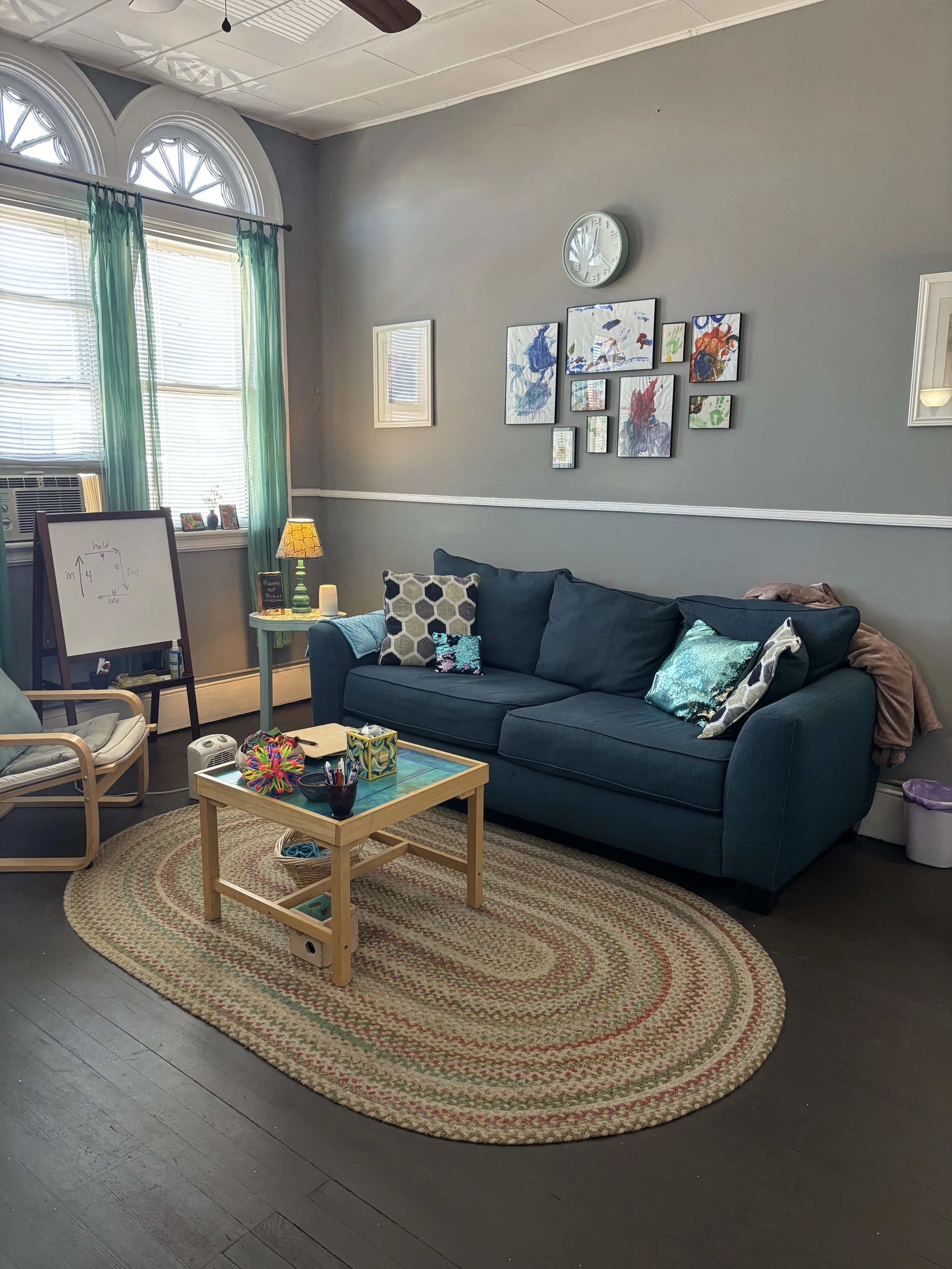 Living room with a gray wall, a dark blue sofa with decorative pillows, a wooden coffee table with toys and art supplies, a white armchair, a window with green curtains, a round braided rug, a small side table with a lamp and candles, and a wall deco