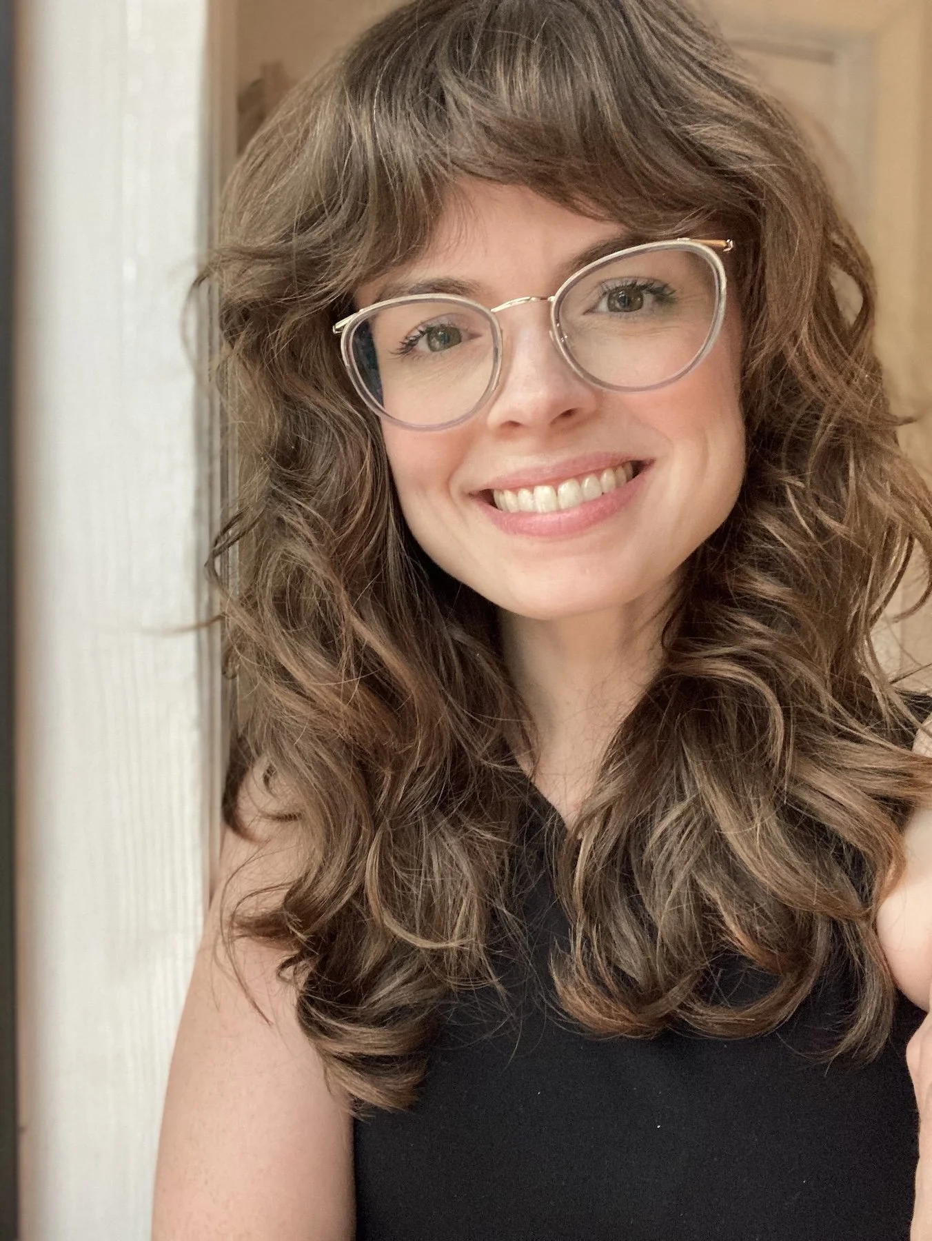 Close-up of a smiling woman with curly brown hair, wearing glasses and a black top.
