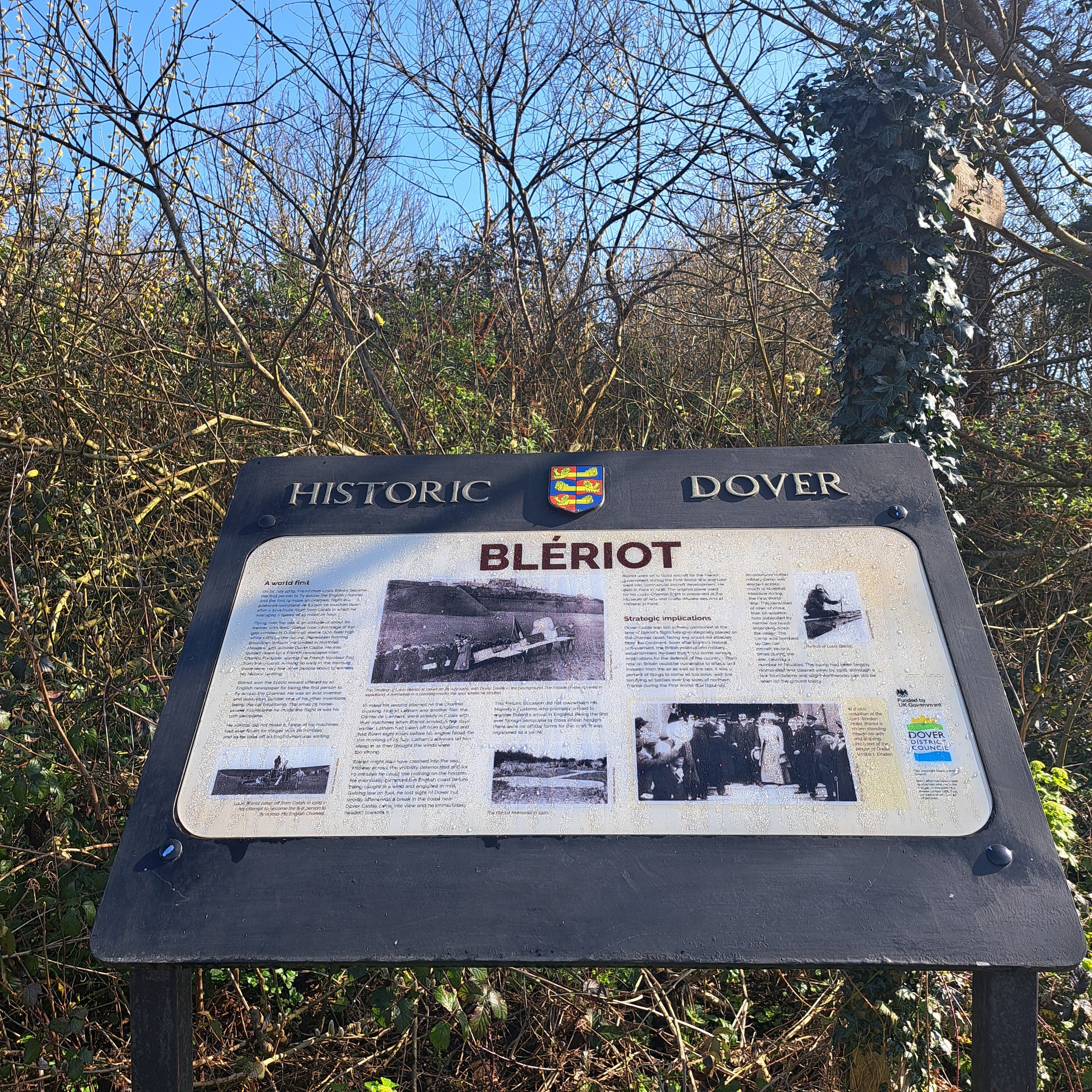 Information board about Blériot in Dover, with historical photos and text, surrounded by bushes and bare trees under a clear blue sky.