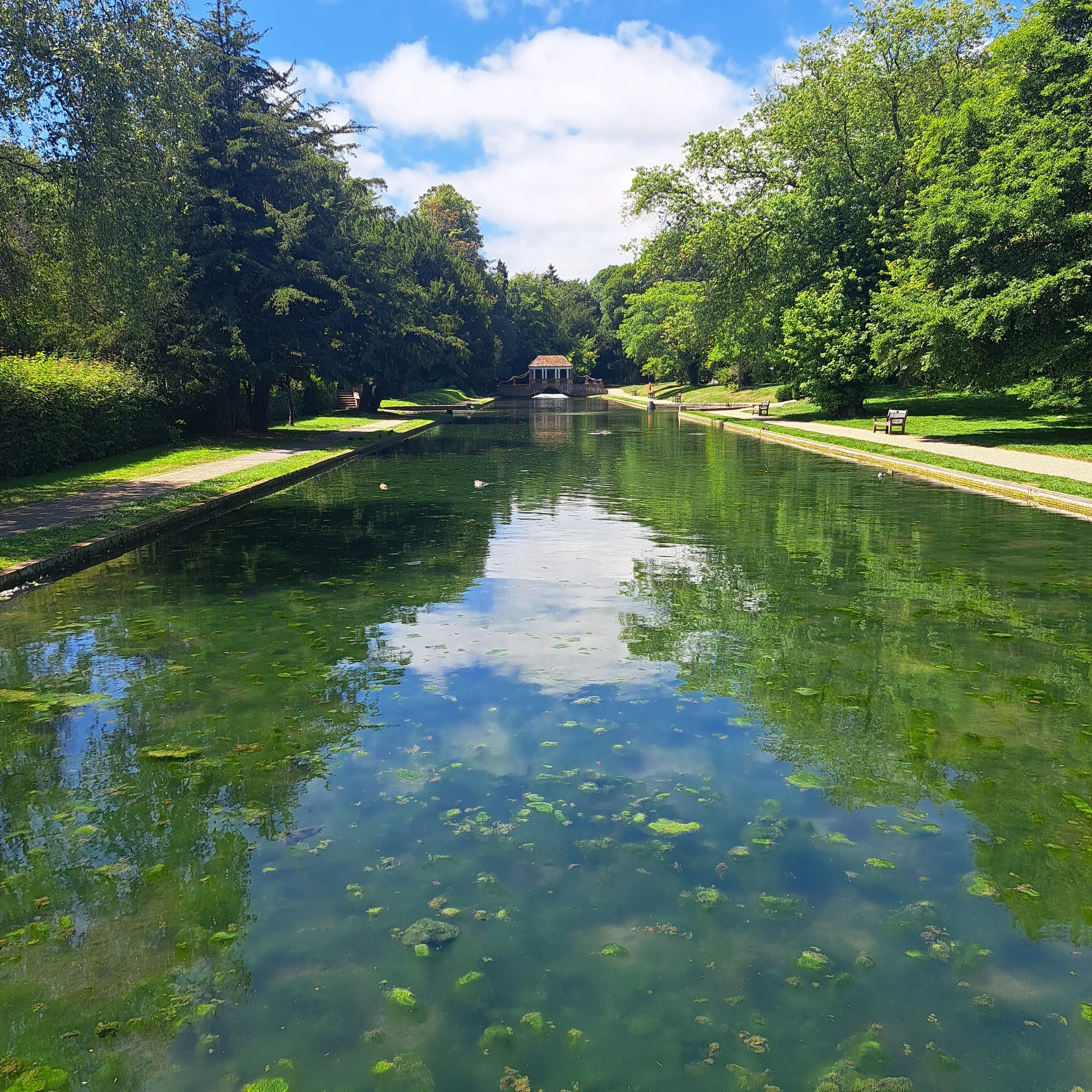A serene park with a long rectangular pond reflecting the sky and trees, with a small building at the far end, green trees on both sides, and a walking path alongside the pond.