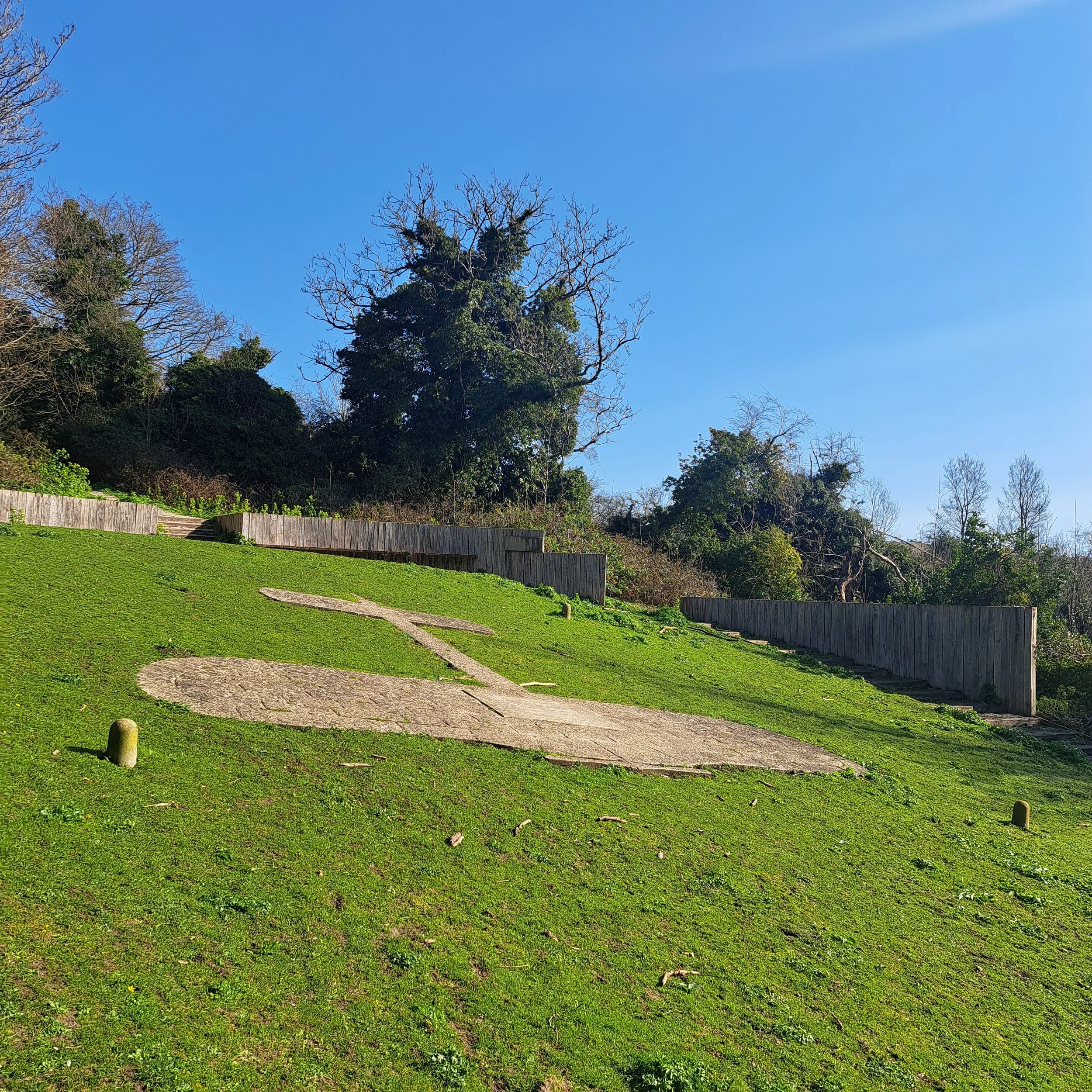 A grassy hillside with a stone pathway shaped like a ship, bordered by wooden fences, and trees and bushes in the background under a clear blue sky.