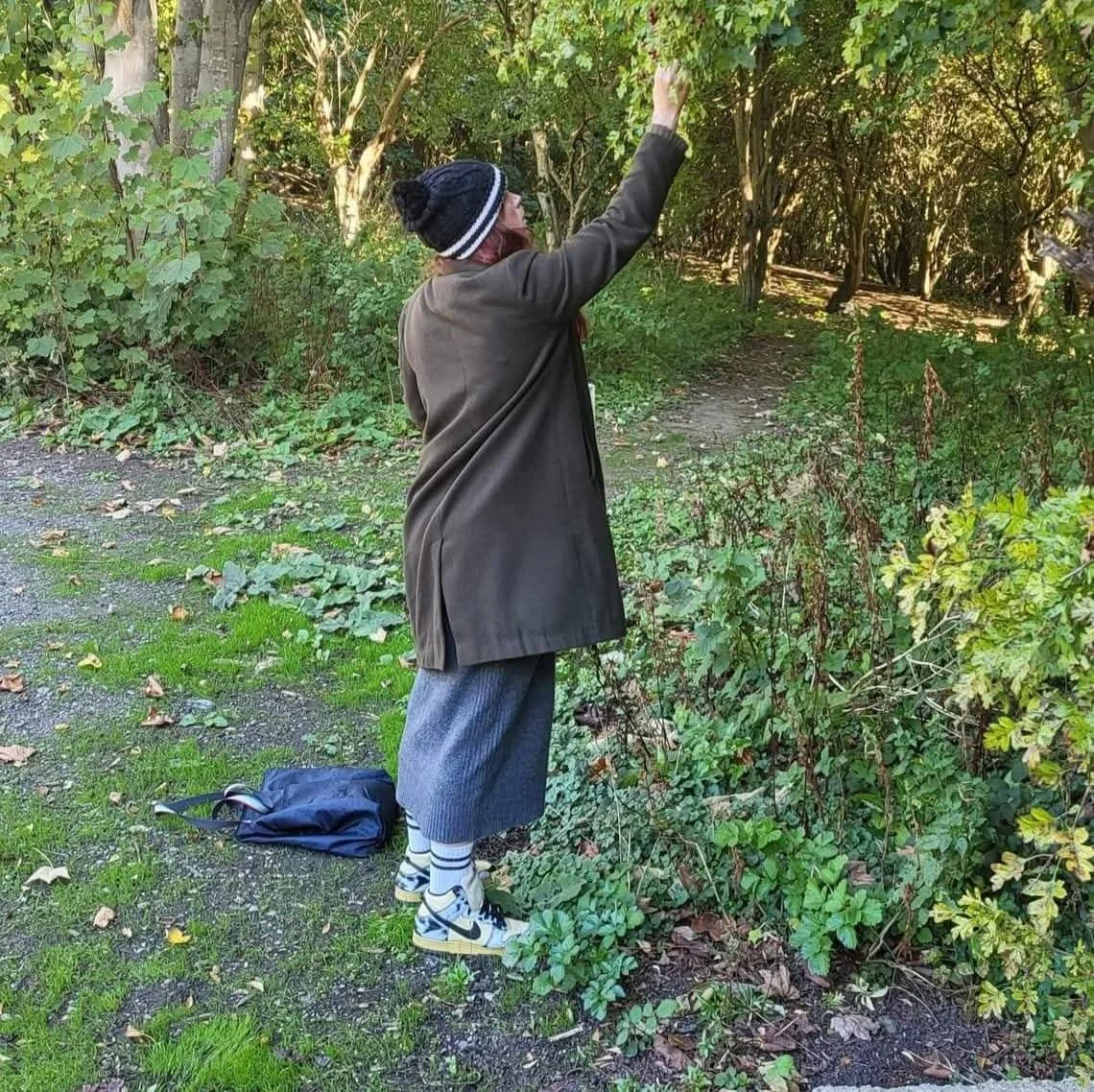 Person in a winter coat, beanie, sneakers, and facemask standing by bushes on a nature trail, reaching up to touch the tree leaves.