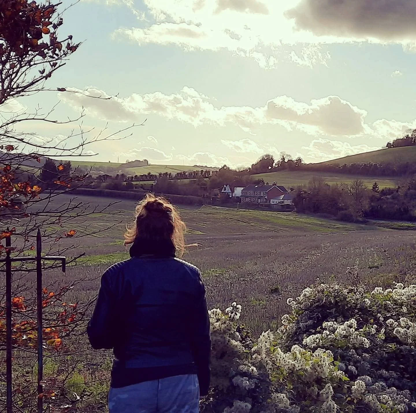 A woman with curly hair in a jacket stands in a rural landscape, overlooking open fields, a house, and rolling hills under a partly cloudy sky.