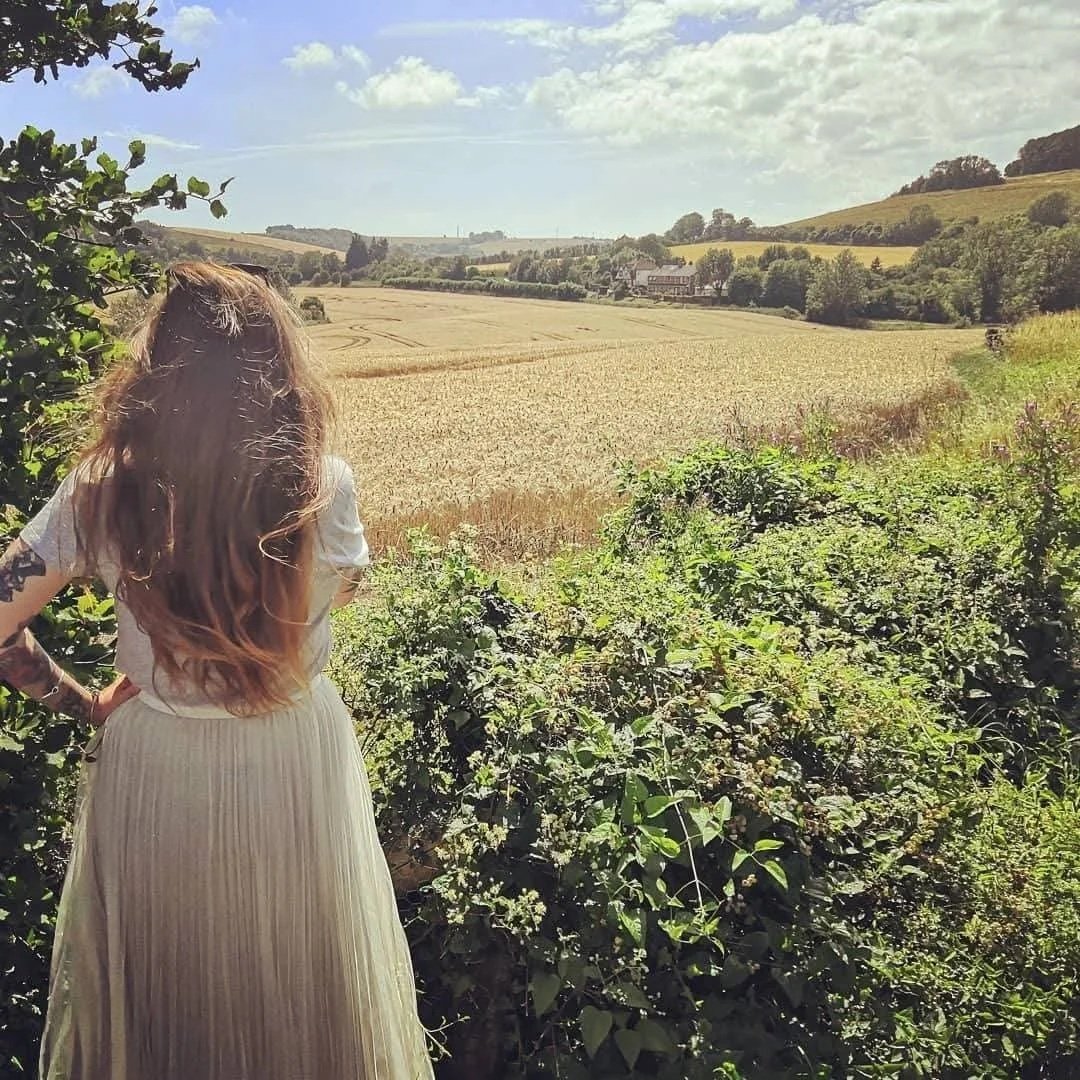 A woman with long hair wearing a white dress standing among green bushes, overlooking a field of crops with houses and rolling hills in the distance under a partly cloudy sky.