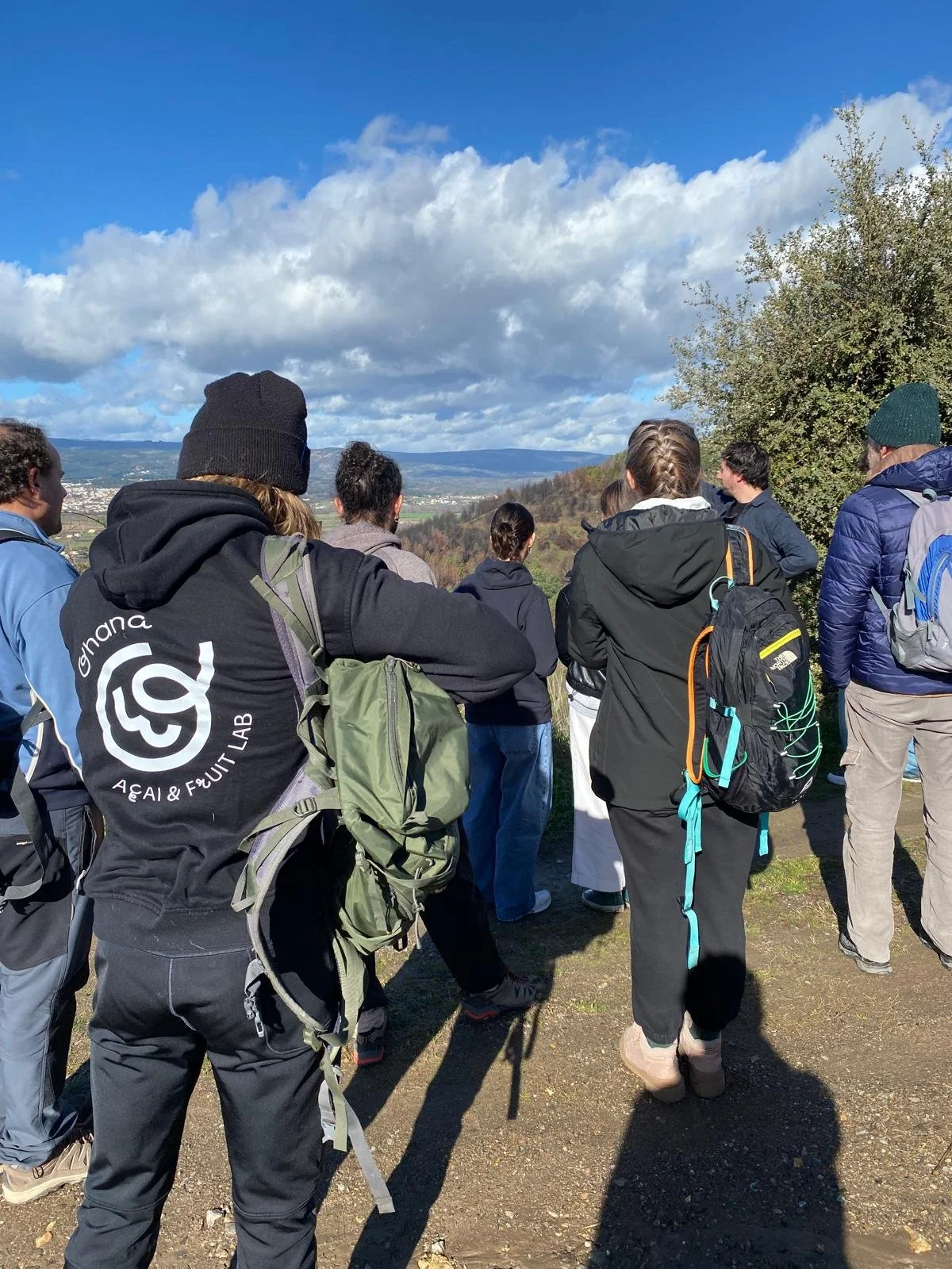 Gruppo di persone in abbigliamento da escursionismo, con zaini, in un paesaggio rurale con cielo nuvoloso.