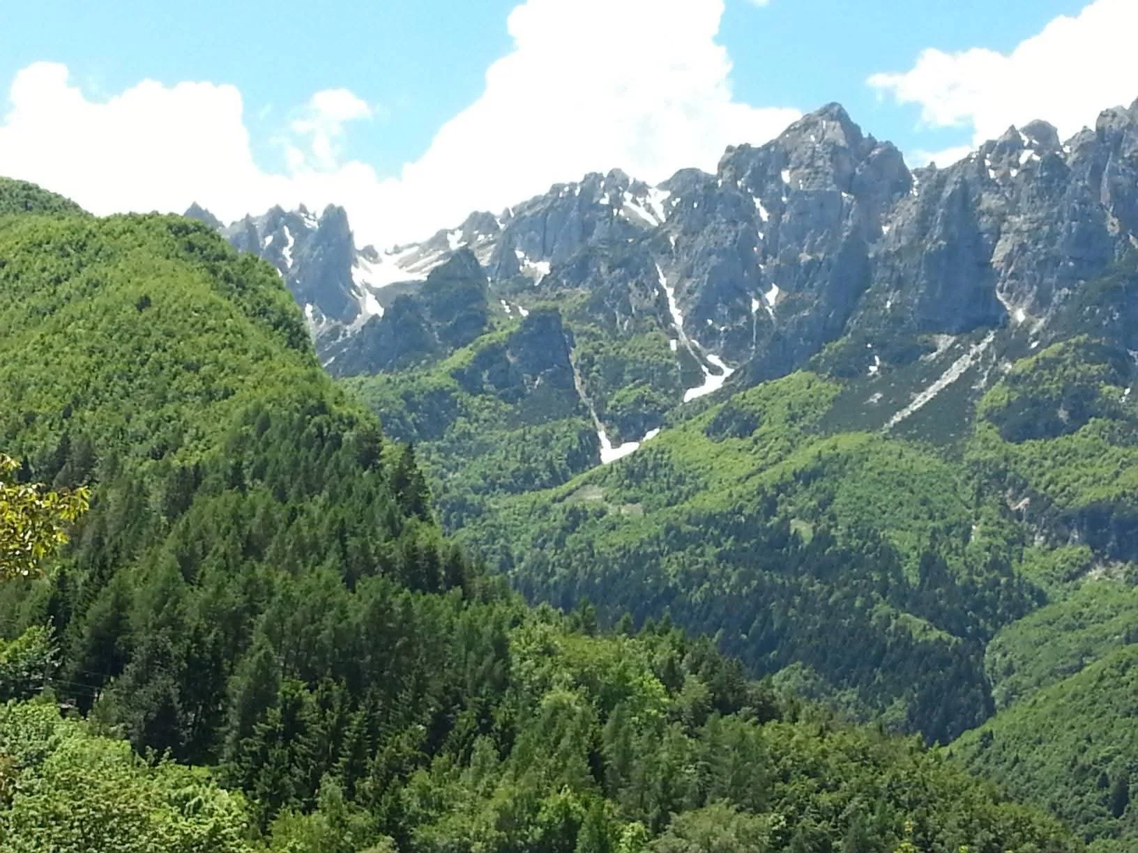 Paesaggio montano con piccole valli ricoperte di foreste e alte cime con neve in lontananza, cielo blu con nuvole