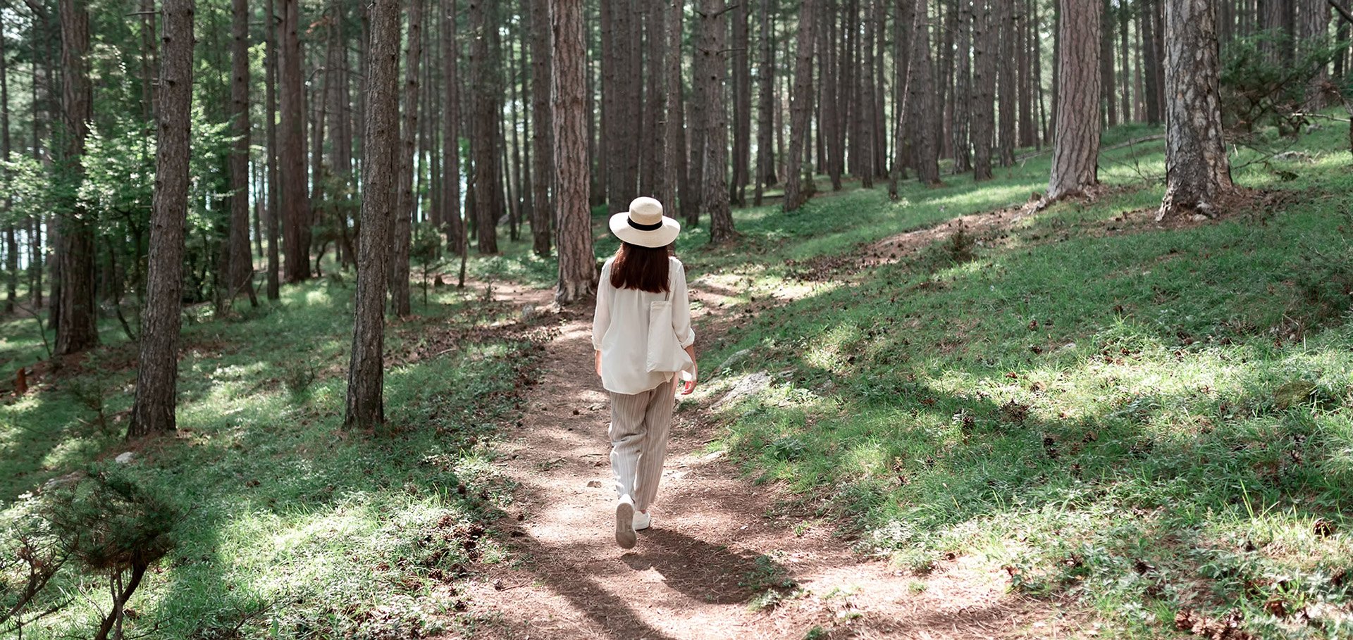 Eine Frau läuft auf einem Waldpfad durch einen Nadelwald, trägt einen weißen Hut und eine weiße Jacke.