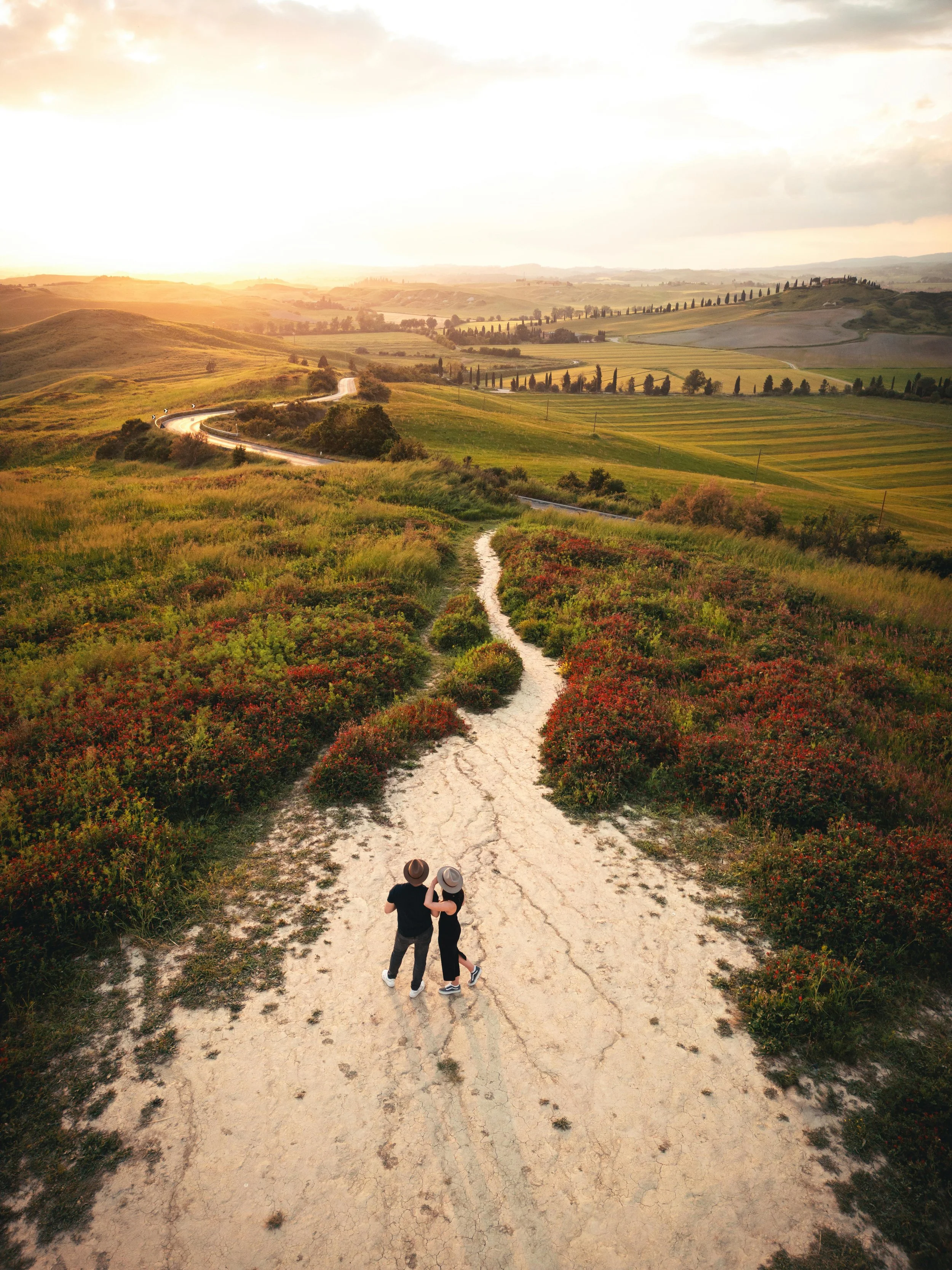 Zwei Menschen gehen Hand in Hand auf einer schmutzigen Landstraße durch eine grüne, hügelige Landschaft bei Sonnenuntergang