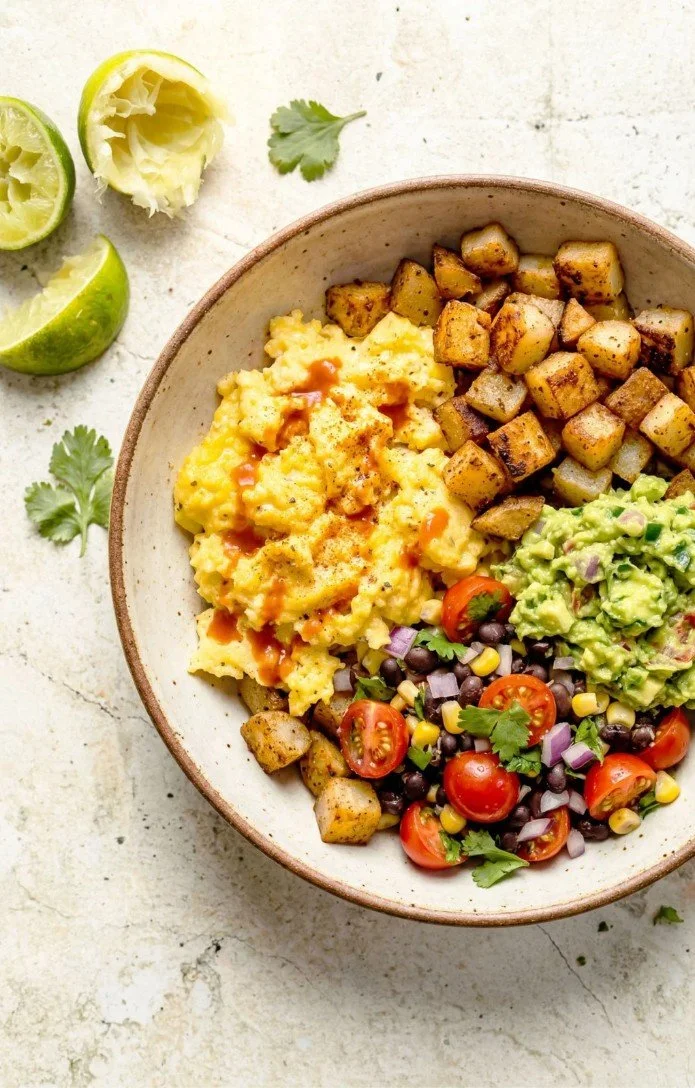A bowl of breakfast with scrambled eggs topped with hot sauce, roasted potatoes, guacamole, and a black bean and cherry tomato salad, garnished with cilantro, with lime wedges, onion, and cilantro around it.