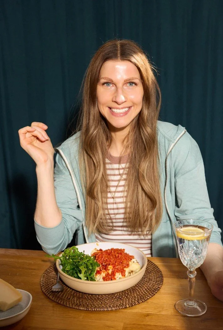 Sabine Venier wearing a light blue jacket over a striped shirt, sitting at a wooden table with a bowl of baked cauliflower topped with tomato sauce and a side of greens, and a glass of water with lemon.