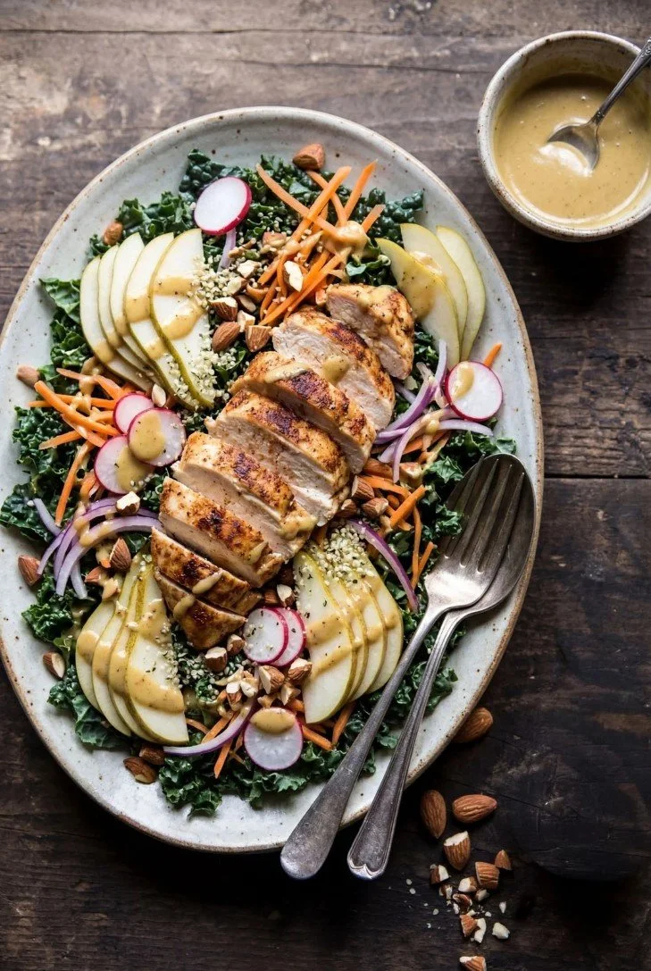 A salad with sliced grilled chicken, sliced apples, radishes, carrots, red onions, chopped nuts, and greens, served with a mustard-based dressing in a small bowl, on a dark wooden surface.
