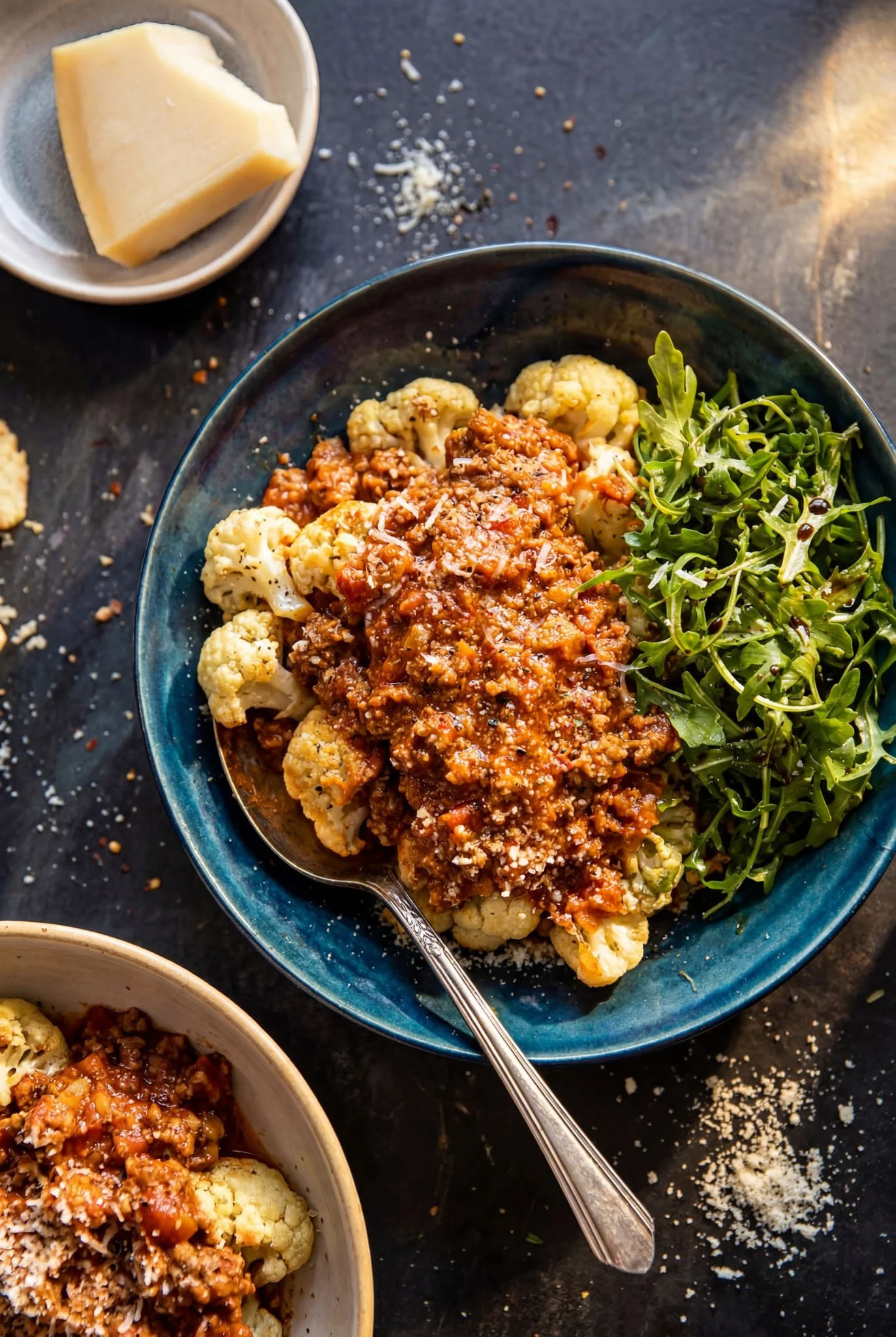 A bowl of roasted cauliflower topped with bolognese sauce and grated parmesan cheese, with a side of fresh arugula salad.