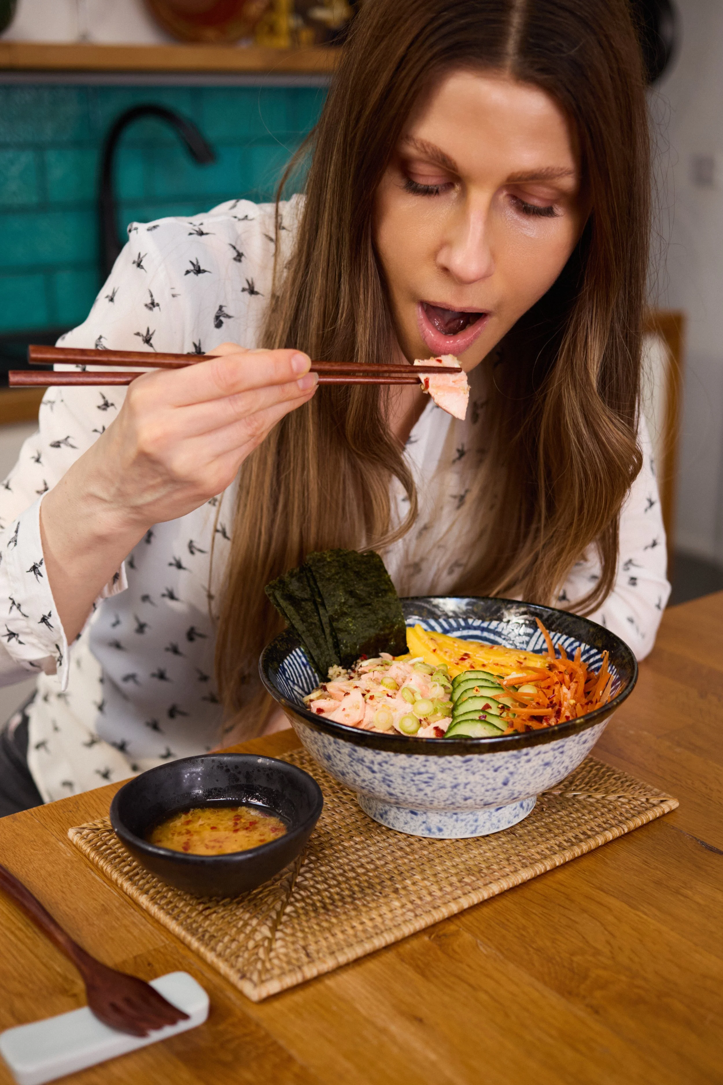 A woman with long brown hair is eating sushi with chopsticks at a restaurant. She is holding a piece of sushi over a colorful bowl of salad that includes cucumber, shredded carrots, and seaweed. A small black dish of soy sauce and a small bowl of dipping sauce are on a woven placemat on a wooden table.