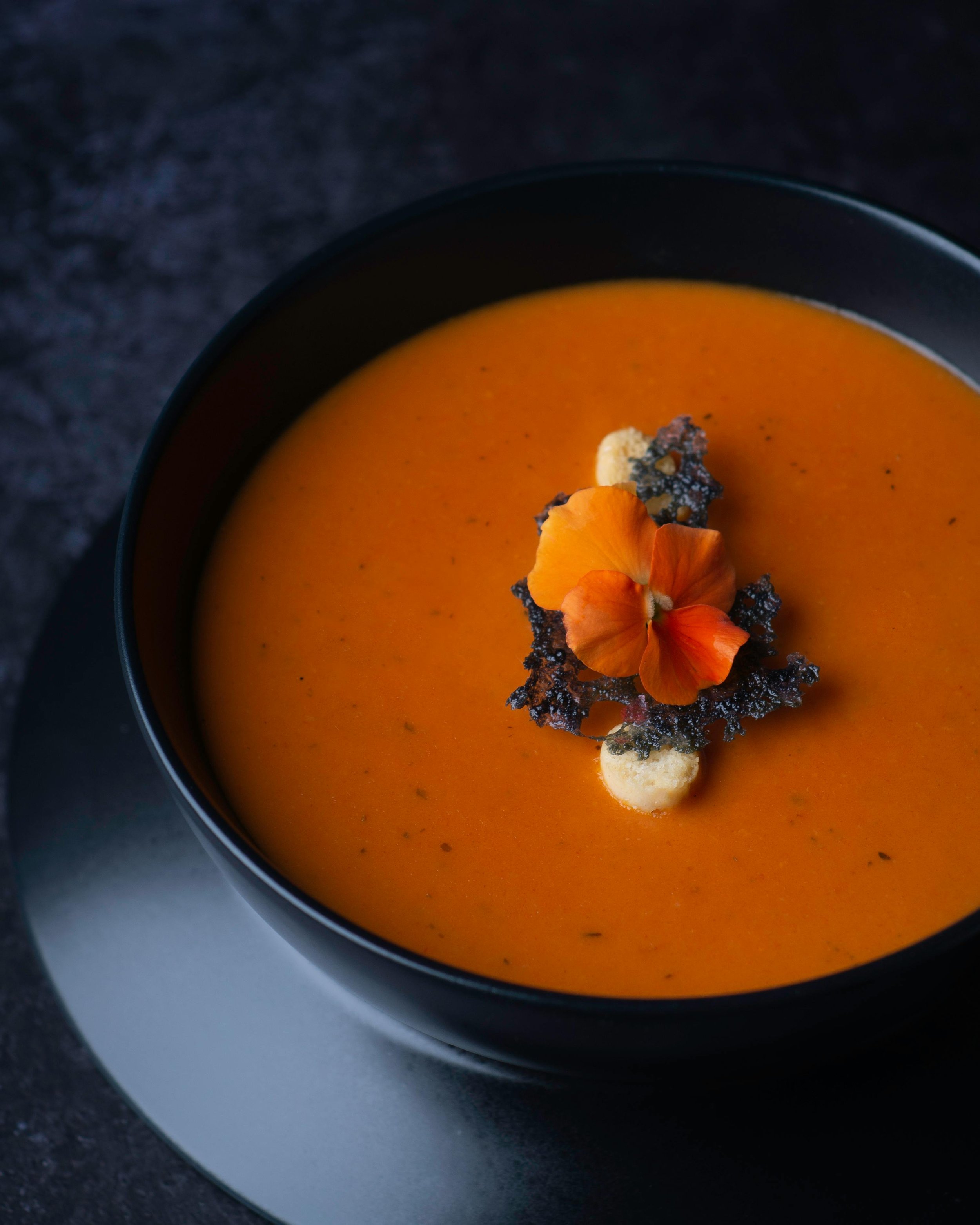 Close-up of a black bowl filled with orange-colored soup, garnished with edible flowers, small white and black crunchy toppings, and black crispy elements, on a white plate on a dark surface.