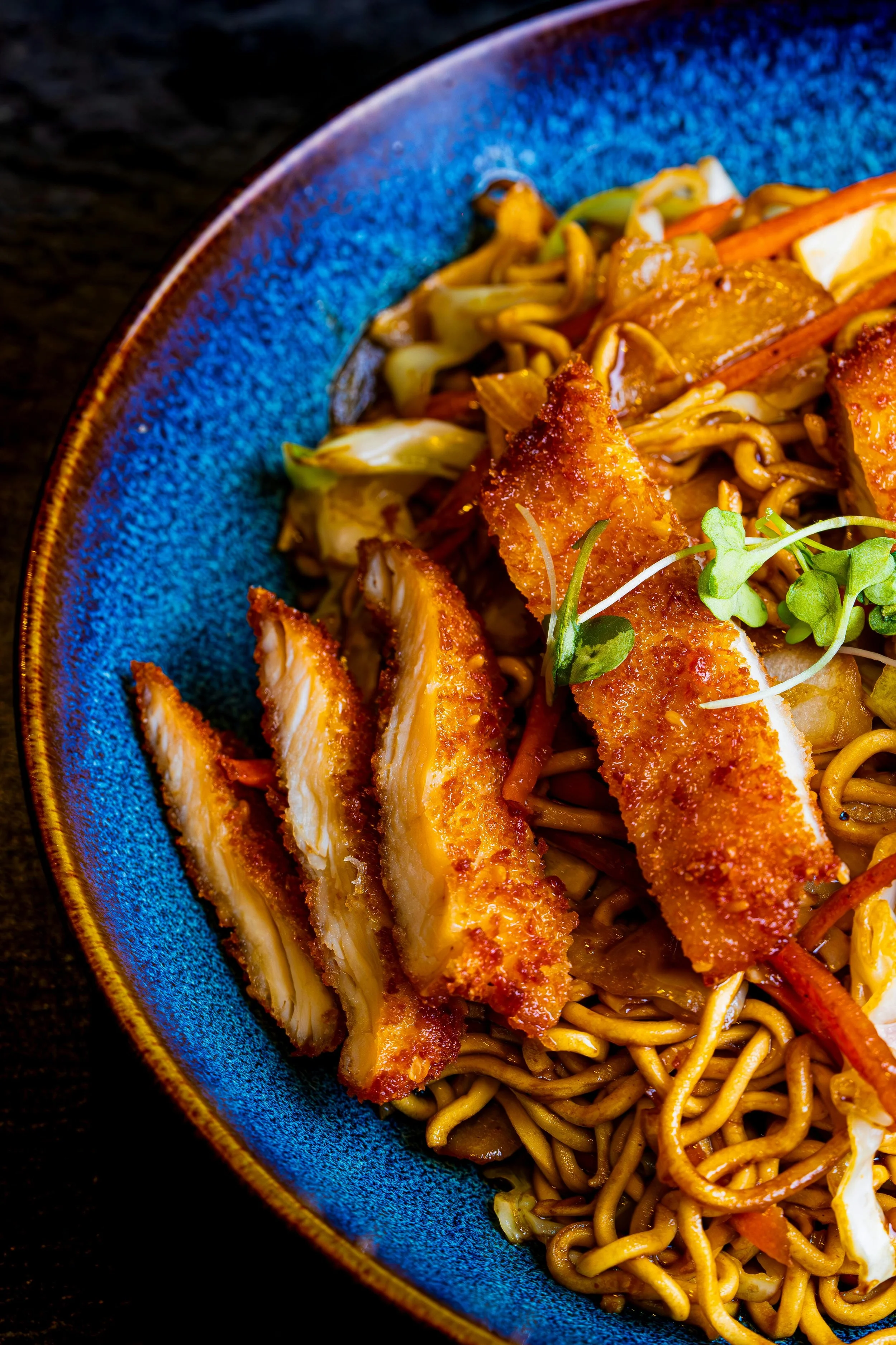 A close-up of a blue ceramic bowl filled with fried chicken strips on top of stir-fried noodles and vegetables garnished with microgreens.