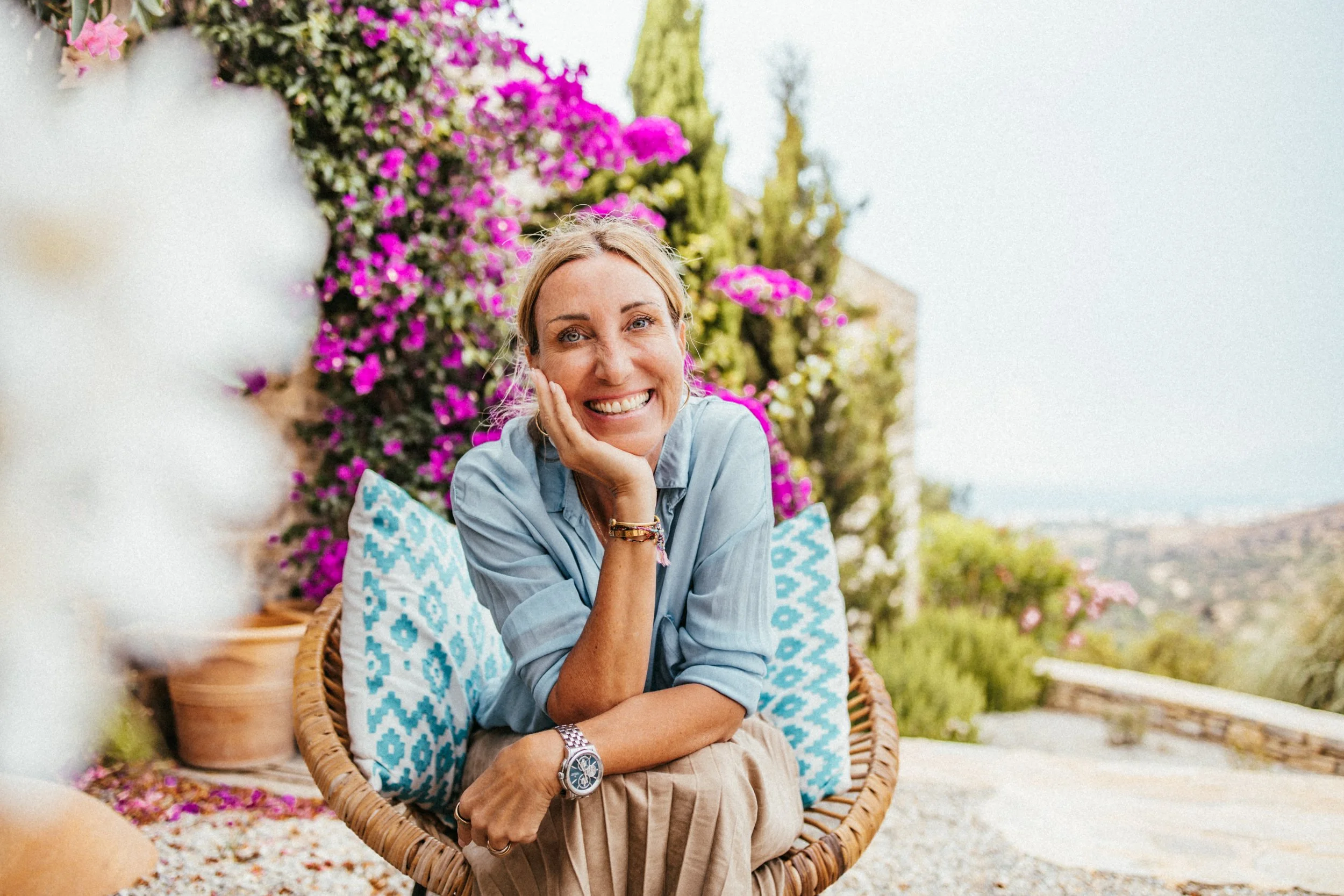 Eine lachende Frau sitzt in einem Korb mit blauen Kissen im Garten mit pinken und weißen Blumen im Hintergrund.